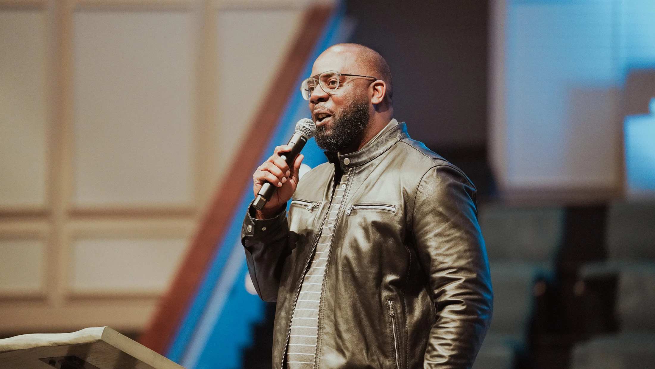A man with glasses and a beard, wearing a black leather jacket, stands on stage and speaks into a microphone with a blurred background.