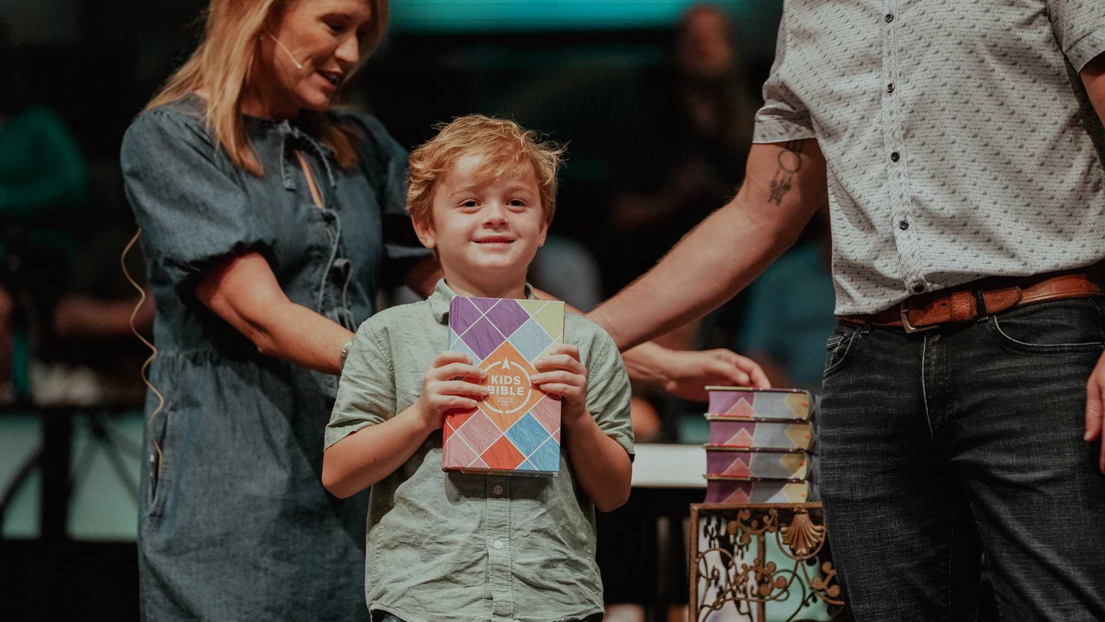 A smiling young boy stands on stage with his parents, proudly holding a new colorful book labeled 'Kids Bible'.