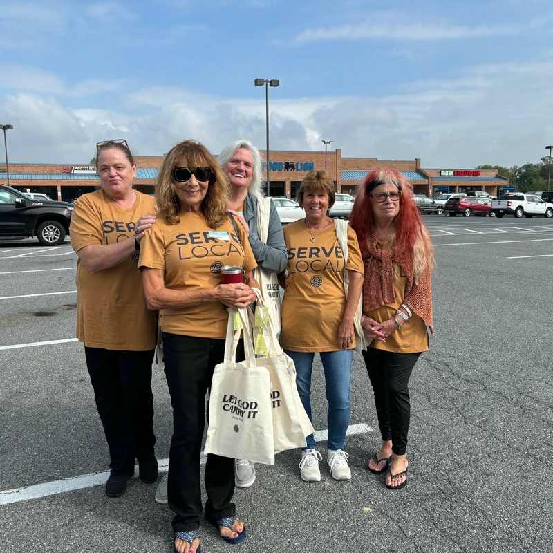 Five women in 'Serve Local' T-shirts smile together in a Milton, Delaware shopping center parking lot during The Crossing's community service day.
