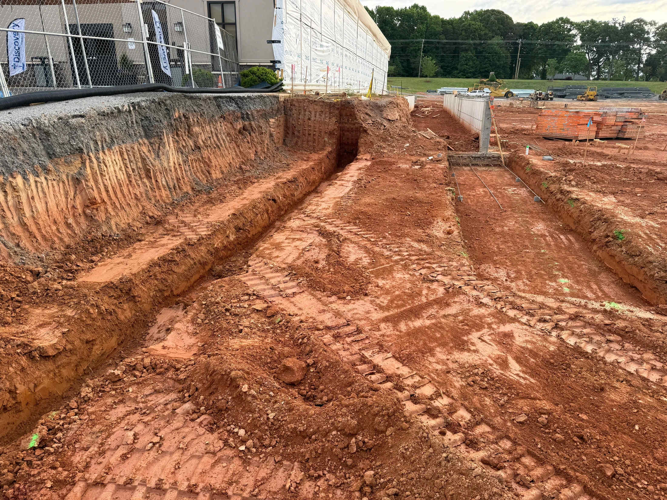 A wide view of a construction site with deep trenches dug into red clay soil. A building under construction is in the background behind a chain-link fence.