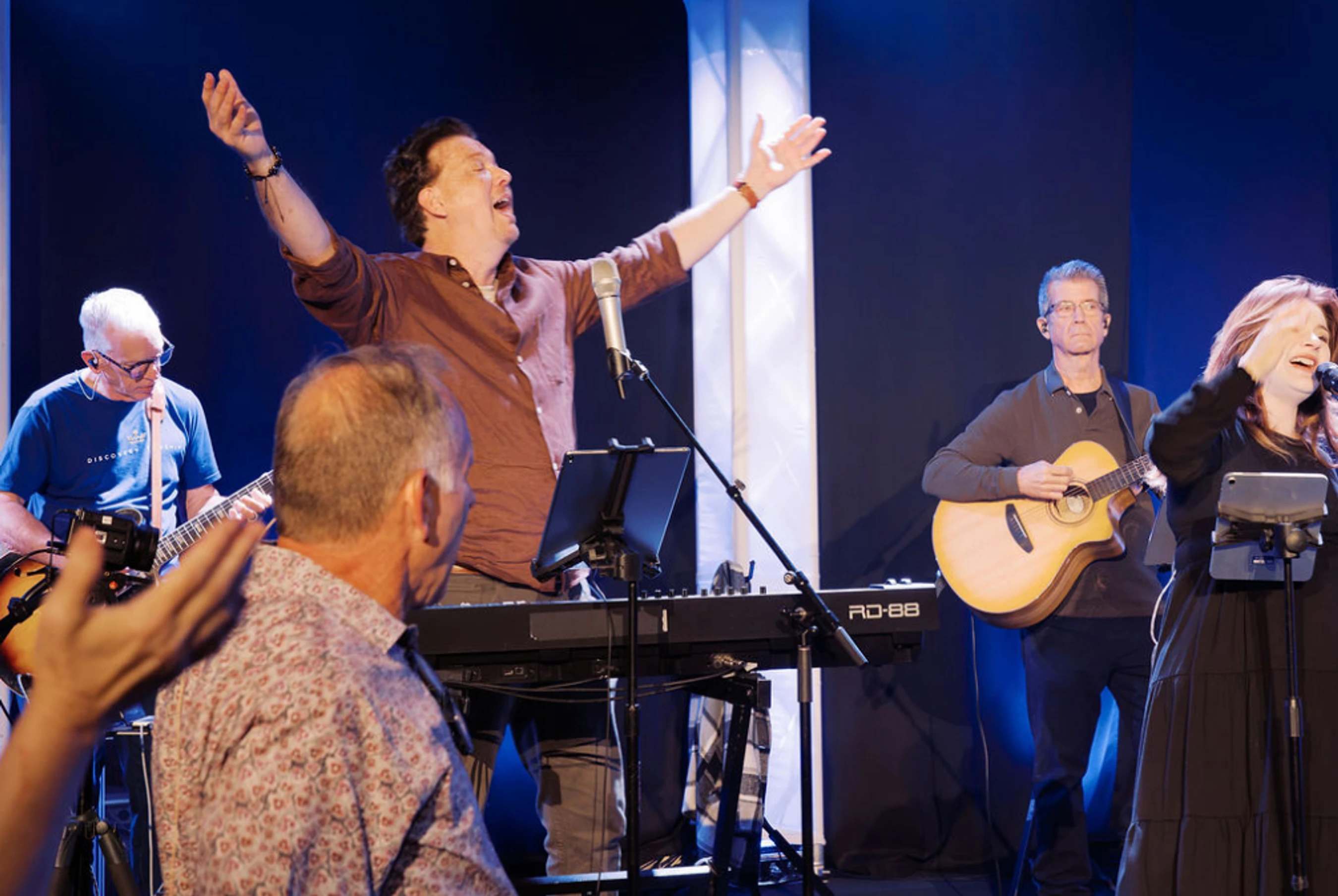 A band performs on stage. A male singer with arms outstretched stands at a keyboard, flanked by guitarists and another singer, all under blue stage lights.