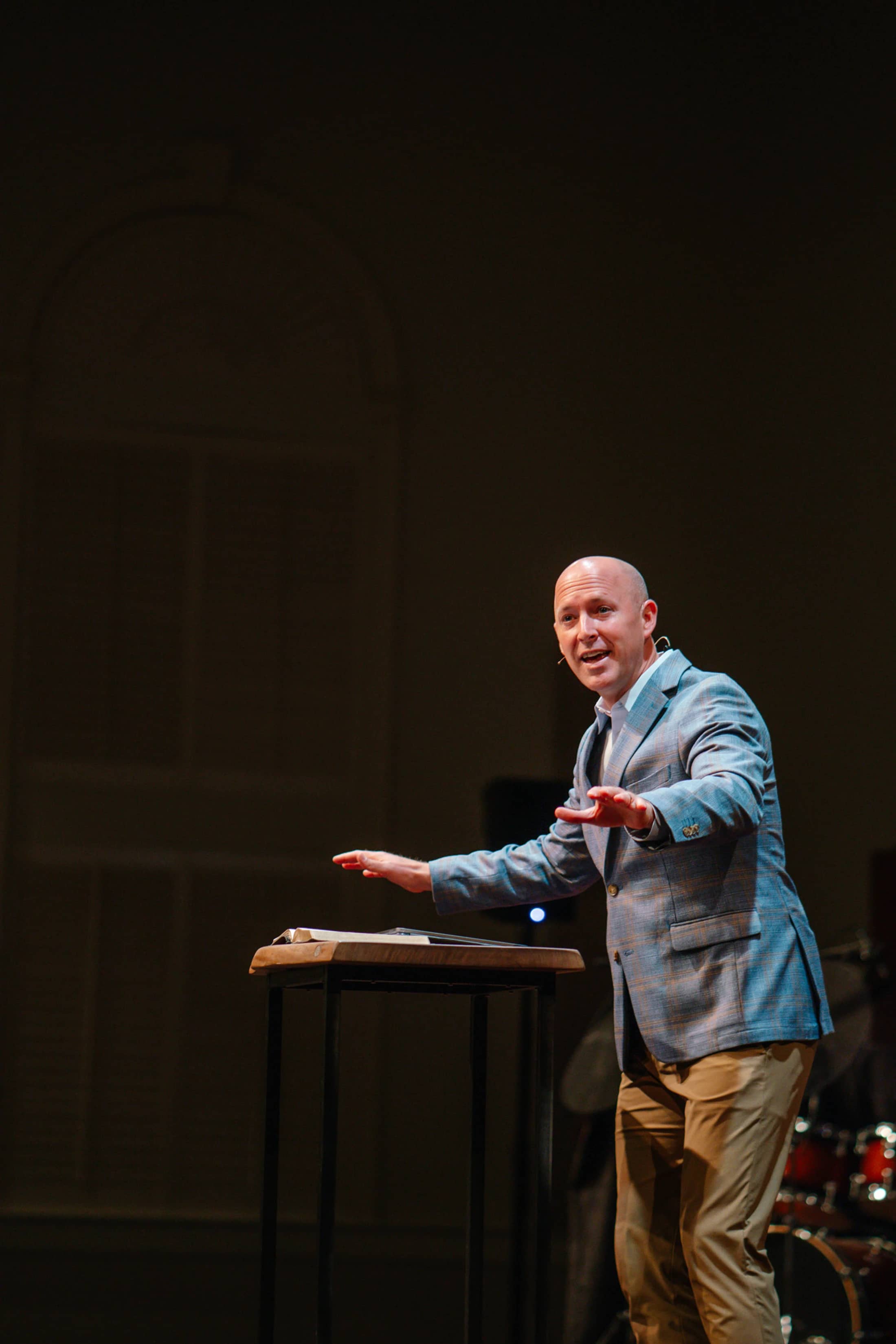 A bald man wearing a blue plaid blazer and khaki pants speaks enthusiastically from behind a lectern on a dimly lit stage.
