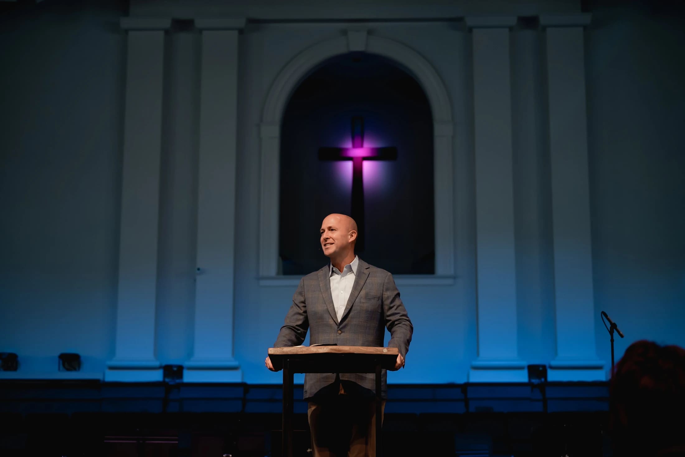 A smiling bald man in a grey blazer stands at a wooden podium on a stage. Behind him, a large cross in an arched alcove glows with purple light.