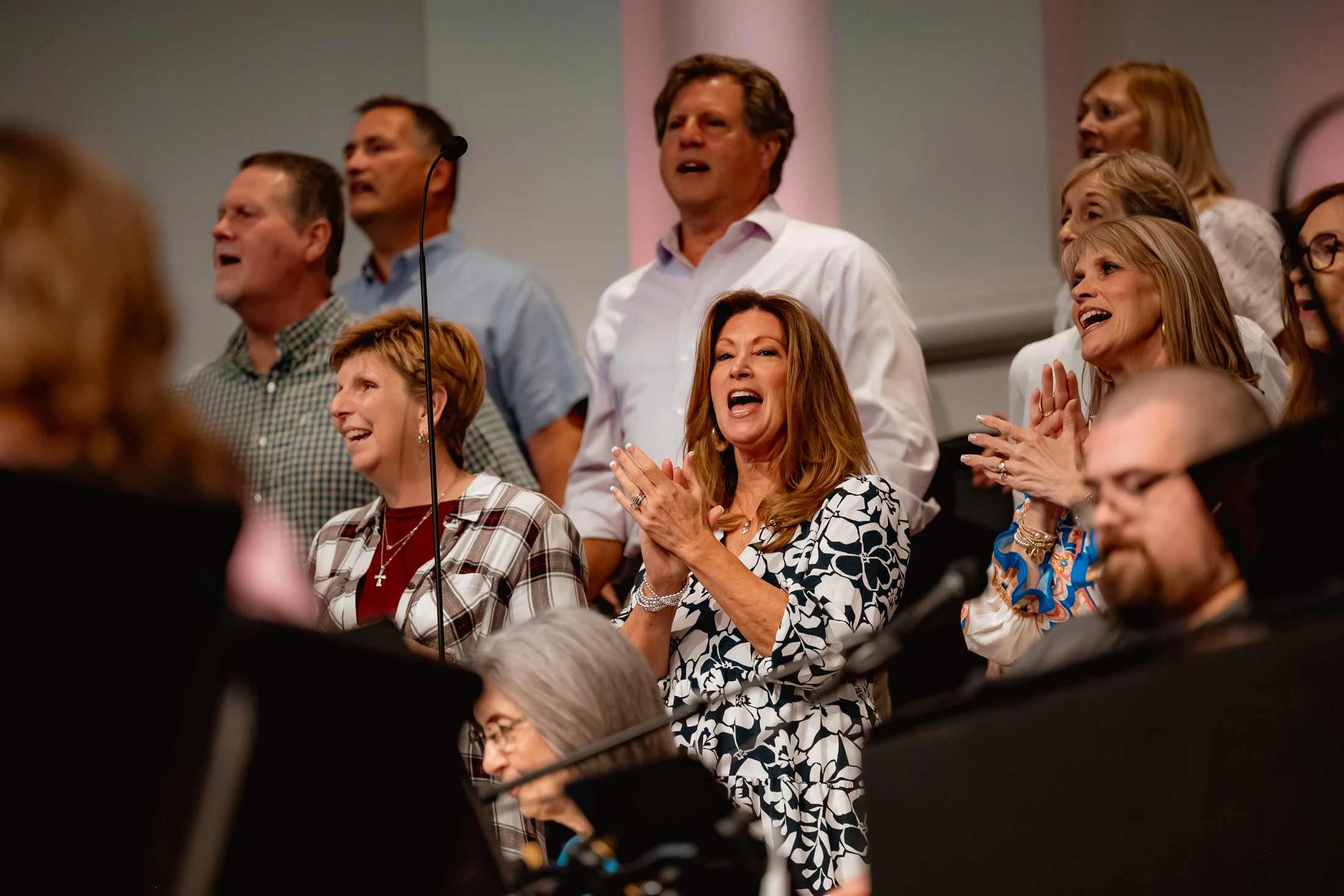 A group of adults in a choir singing joyfully. A woman in a black and white floral dress is in the center, clapping her hands and singing enthusiastically.