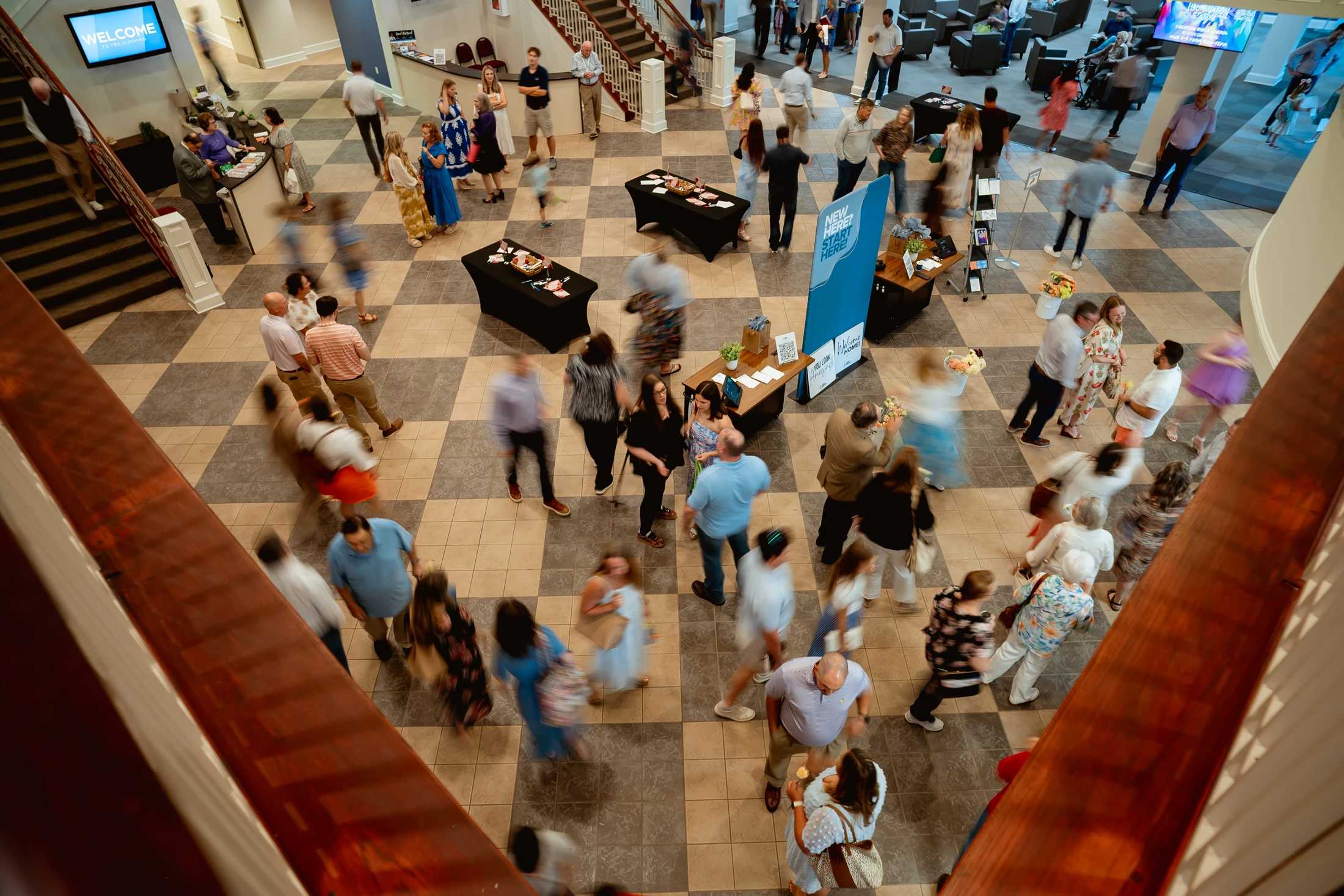 A high-angle view of a bustling church lobby. Many people are blurred with motion as they walk and talk on a checkered tile floor between services.