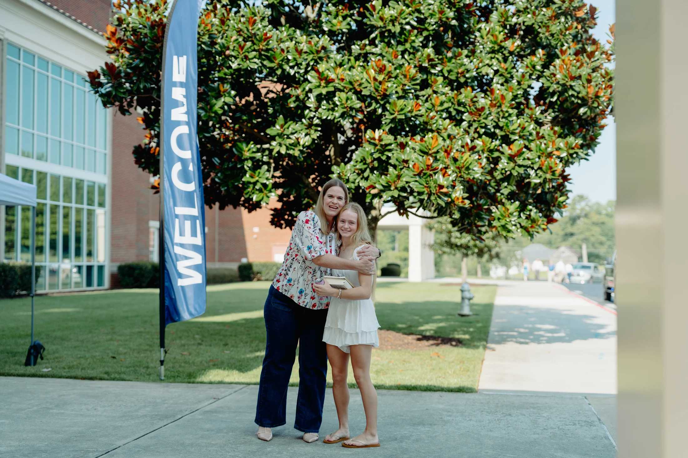 A mother and her teenage daughter share a hug while standing on a sidewalk next to a blue "WELCOME" banner, with a large tree and a building behind them.