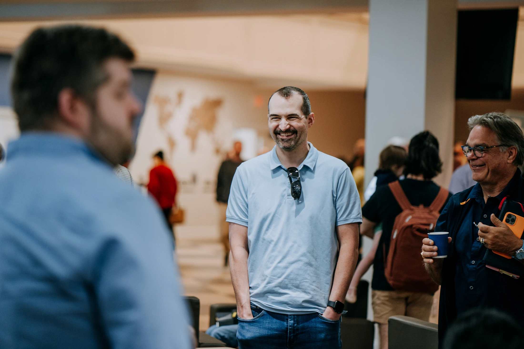 A man in a light blue polo shirt smiles while standing with his hands in his pockets, talking with other people at a casual indoor event.