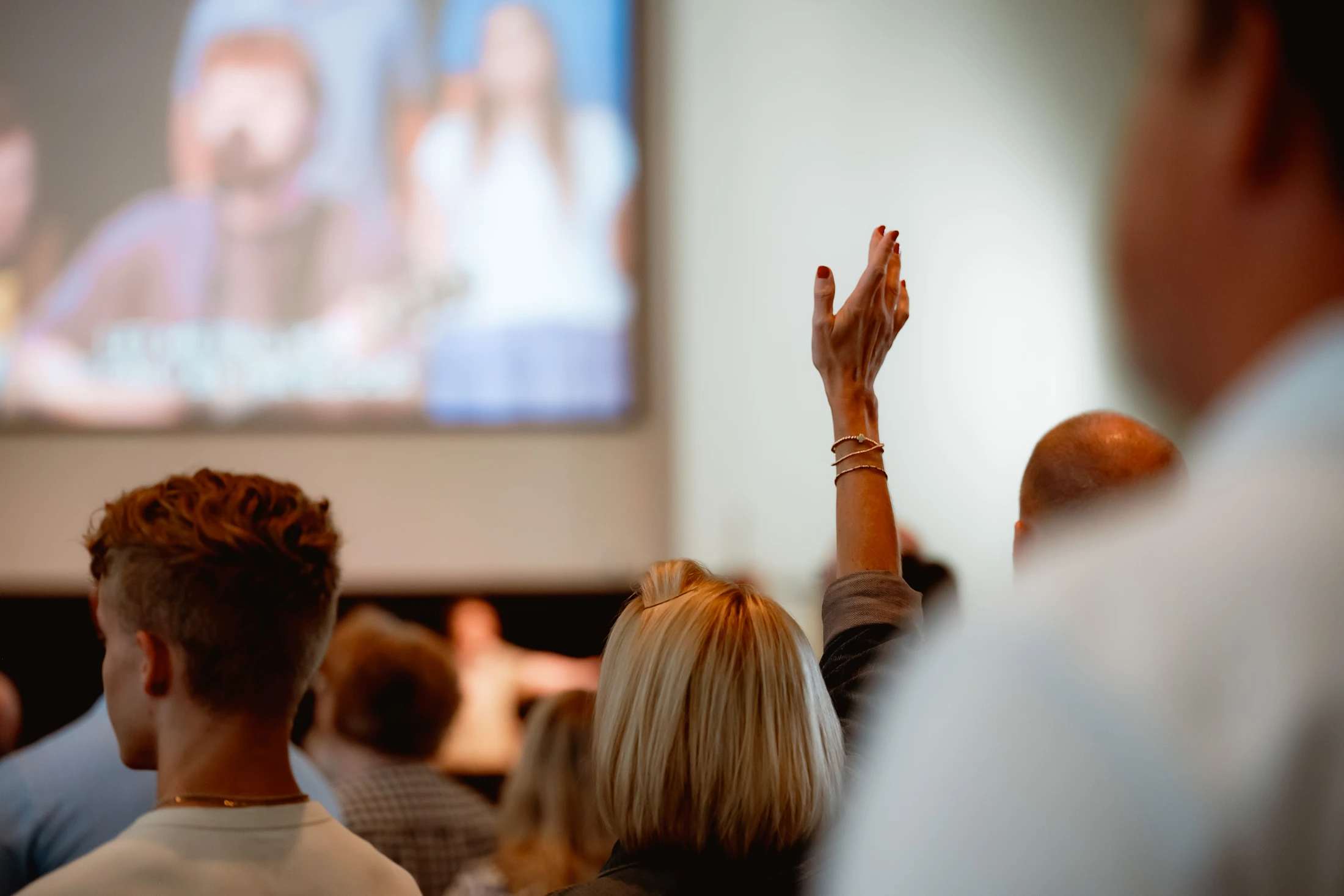 A view from behind a congregation where one person raises their hand in worship. The focus is on the hand, with the stage and screen blurred in the background.