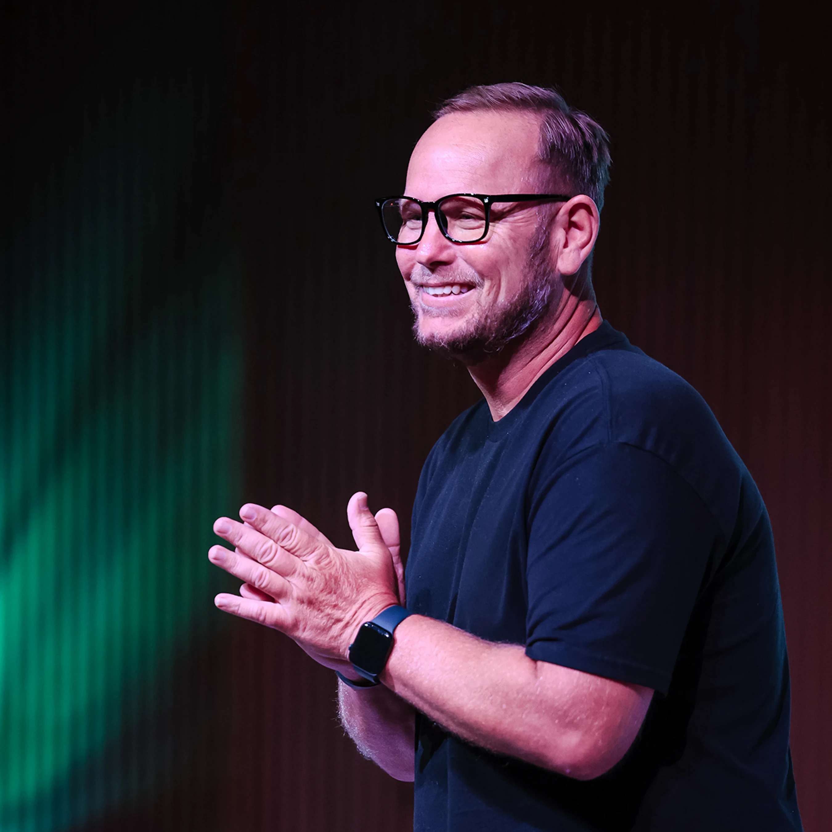 A man with glasses and a black t-shirt smiles as he claps his hands. He is wearing a smartwatch and is lit by green and red stage lights.
