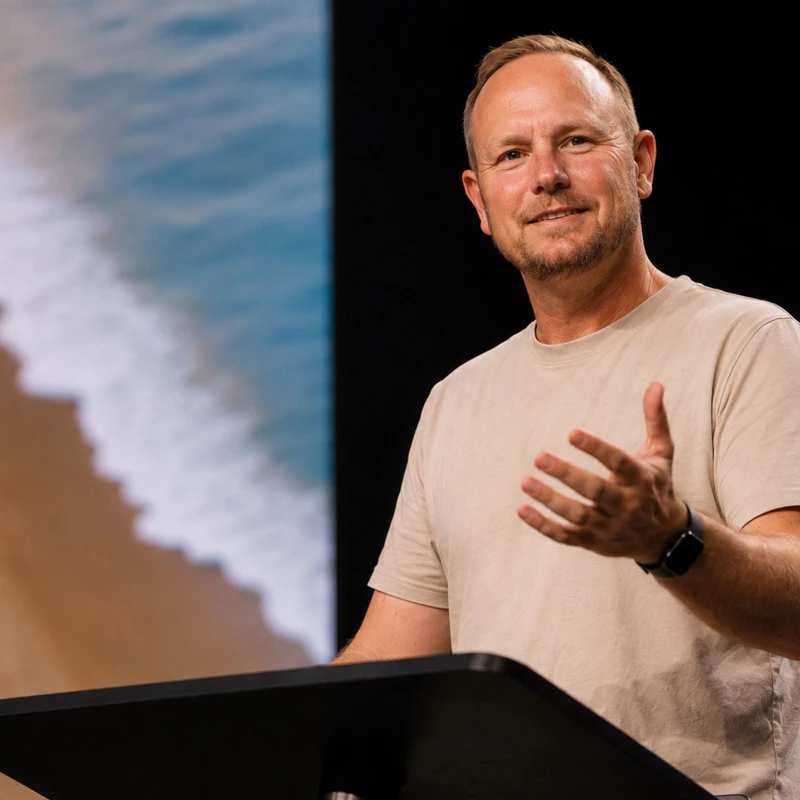 A smiling man in a tan t-shirt stands behind a black podium speaking. A large screen behind him shows an aerial view of waves on a sandy beach.