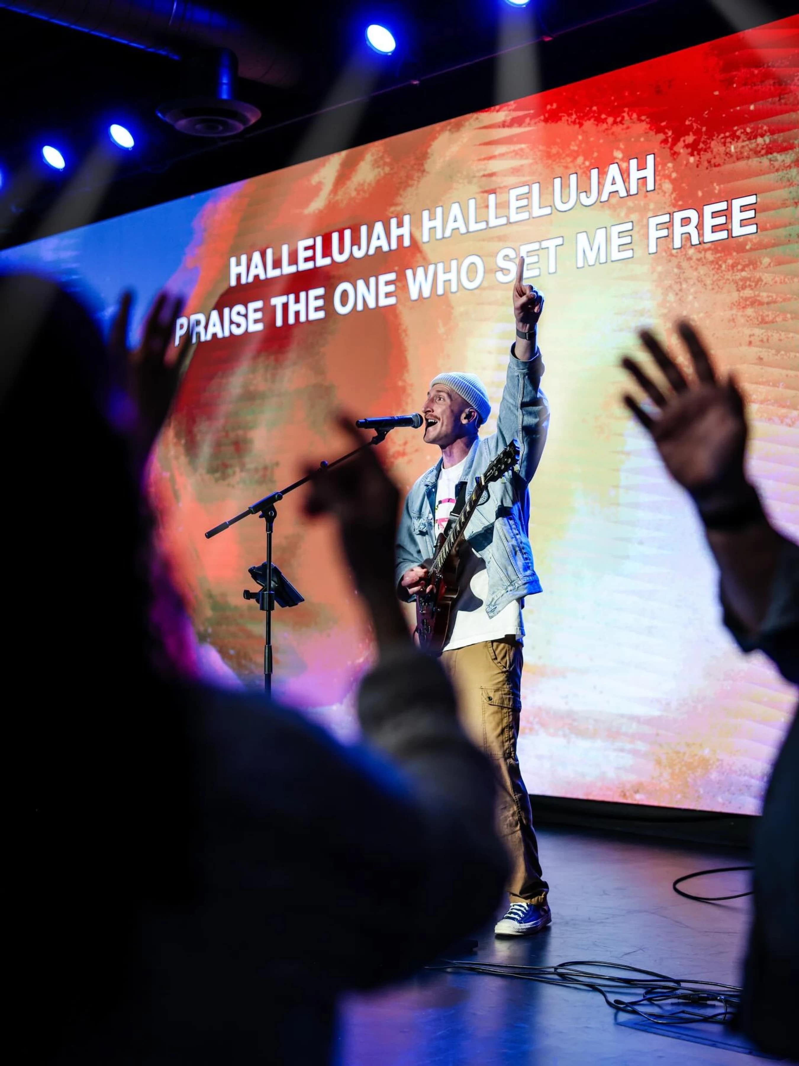 A man on stage sings and plays guitar, pointing up. Behind him, a large screen displays the lyrics, "Hallelujah...Praise the one who set me free."
