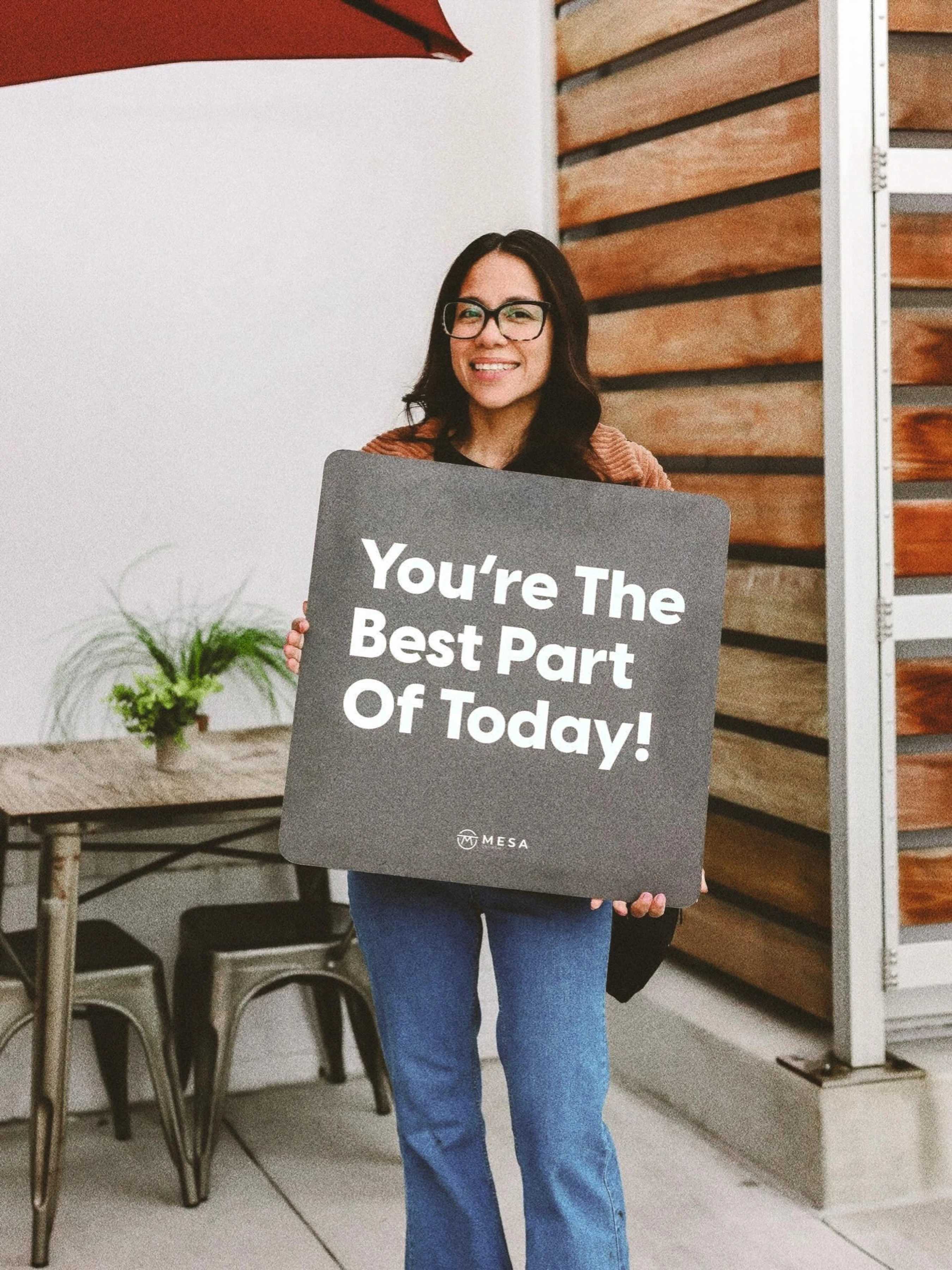 A smiling woman with long dark hair and glasses holds up a gray sign that reads, 'You're The Best Part Of Today!' in white letters.