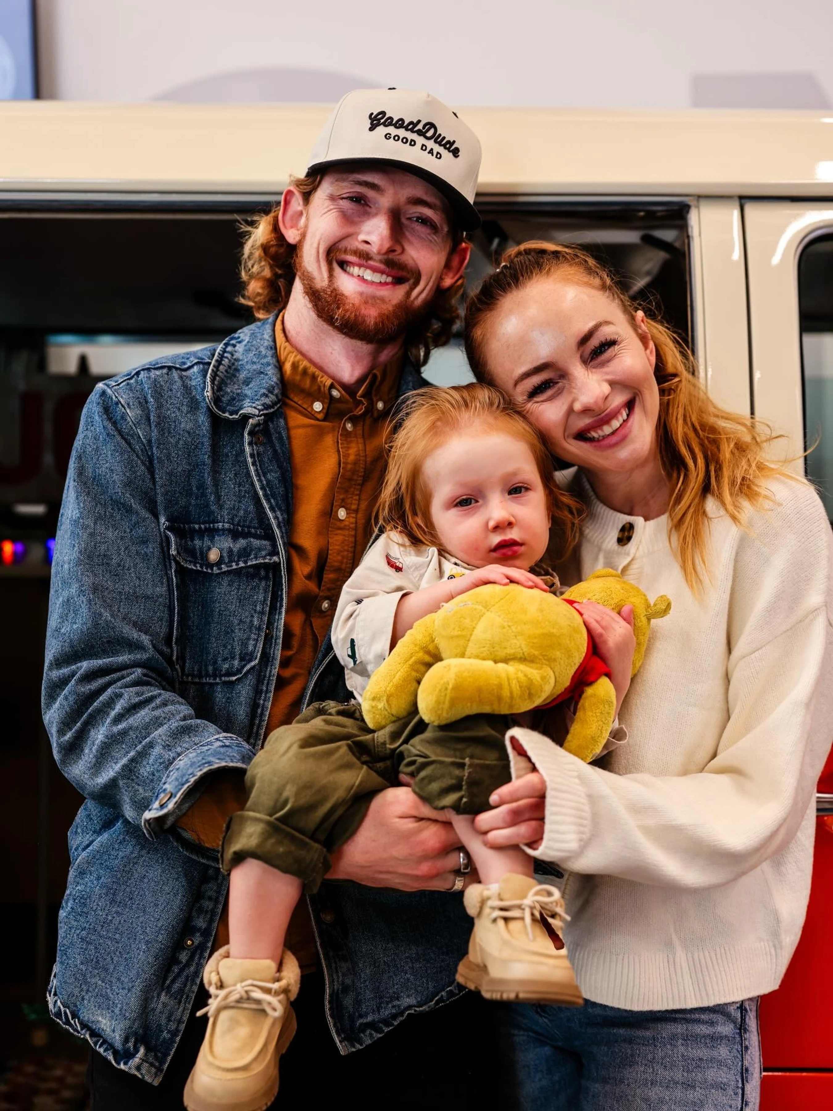 A happy young couple smiles while holding their toddler. The man wears a 'Good Dad' hat, and the child holds a Winnie the Pooh stuffed animal.