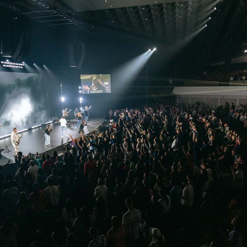 A high-angle view of a large crowd in a dark auditorium, with hands raised towards a band performing on a brightly lit stage during a worship service.