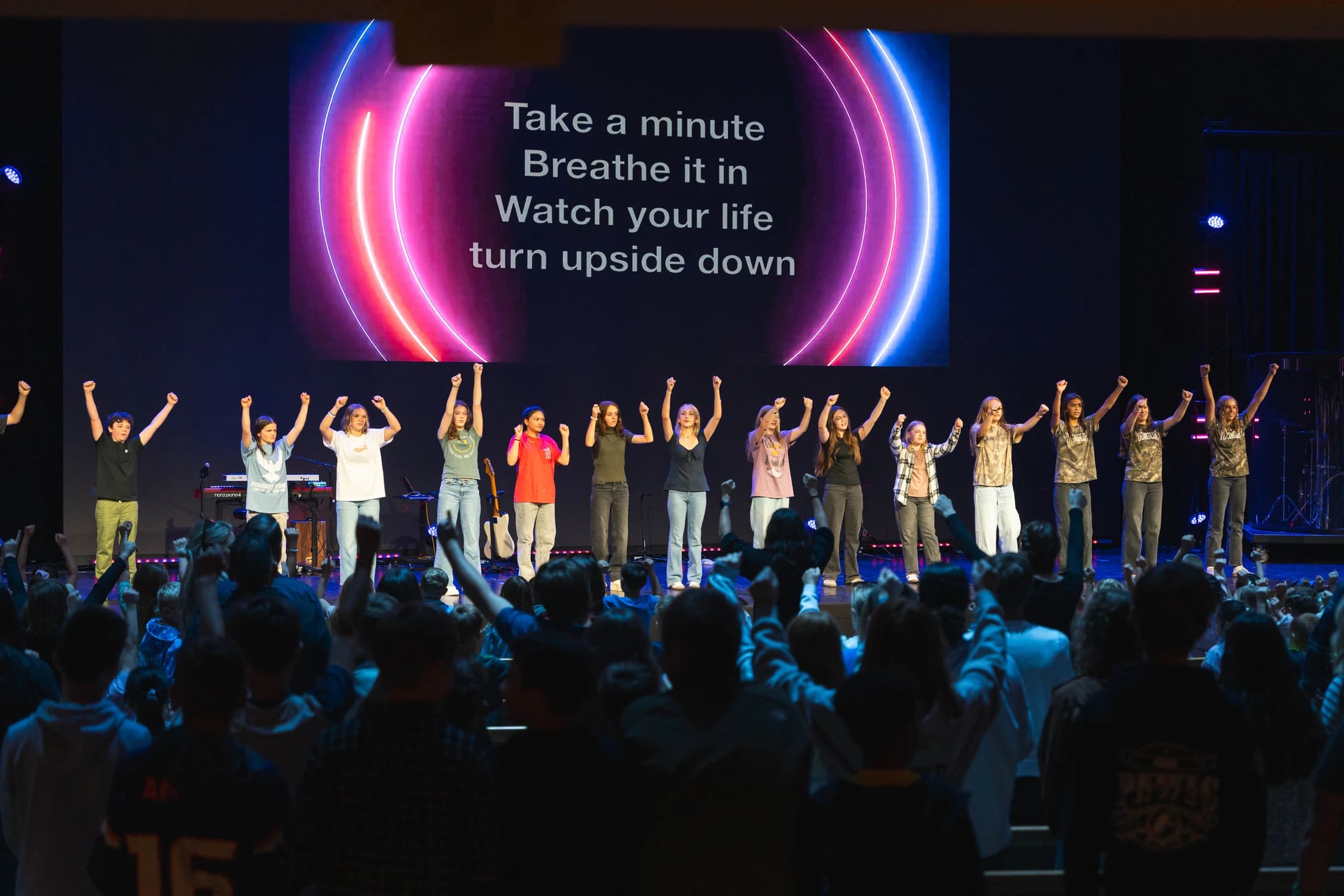 A group of young people stand on a stage with their fists raised, singing in front of a large audience. A screen behind them displays song lyrics.