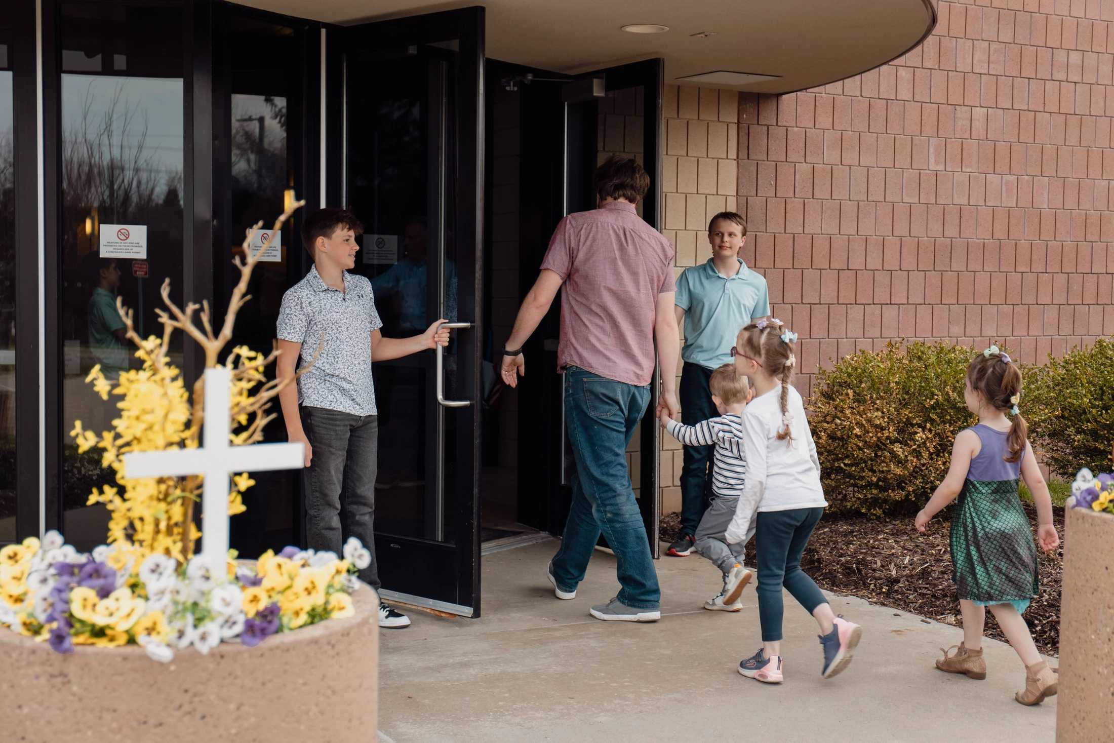A man and five children enter a church building through glass doors held open by a teenage boy. A white cross is visible in a planter in the foreground.