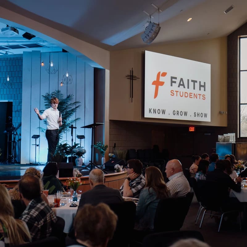 A young man stands on a blue-lit stage, presenting to an audience seated at round tables during a church gathering for Faith Students.
