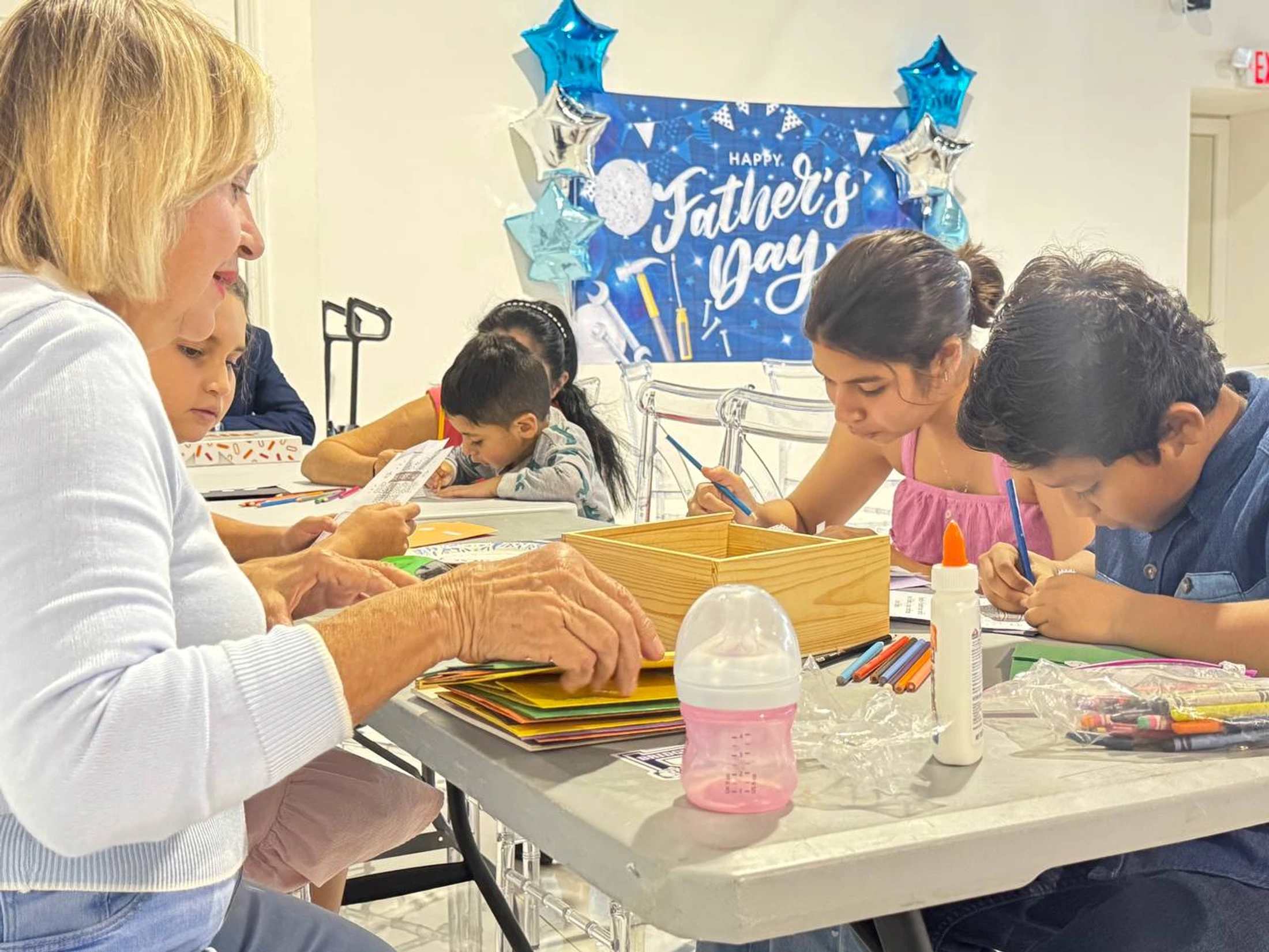 Adults and children sit together at a table, focused on drawing and crafting, with a "Happy Father's Day" banner in the background.
