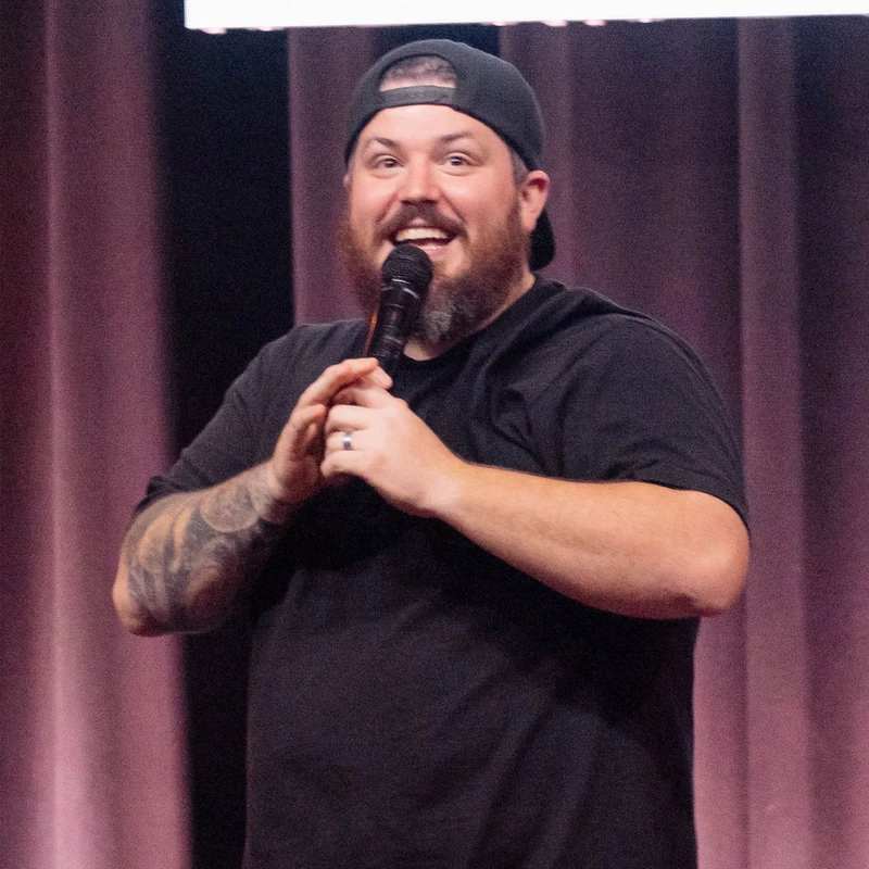 A man with a beard and a backward cap, wearing a black shirt, smiles while speaking into a microphone on stage in front of a purple curtain.