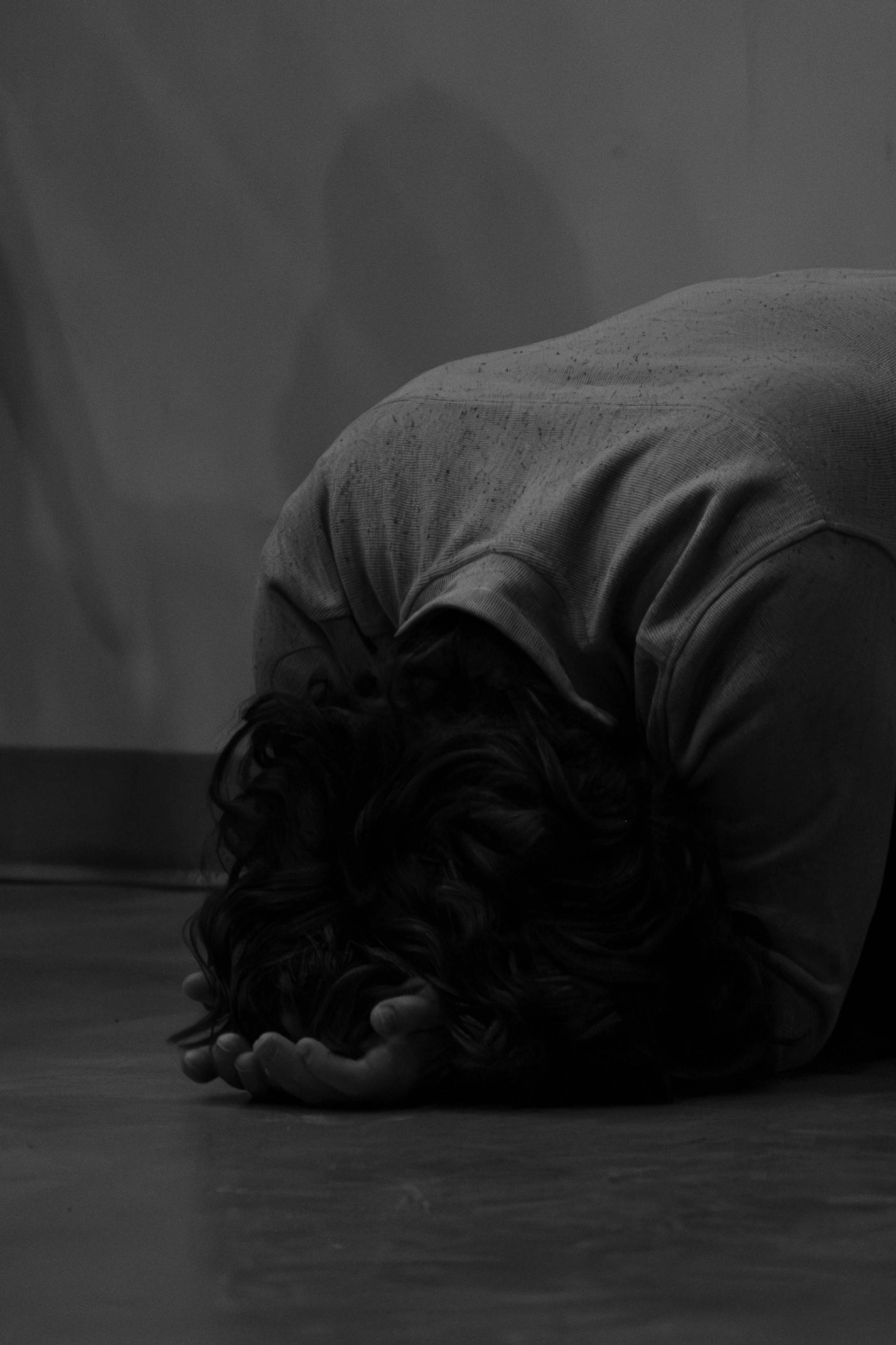 A person with dark hair kneels on the floor, head bowed down to their cupped hands in a black and white image, evoking a sense of sadness or prayer.