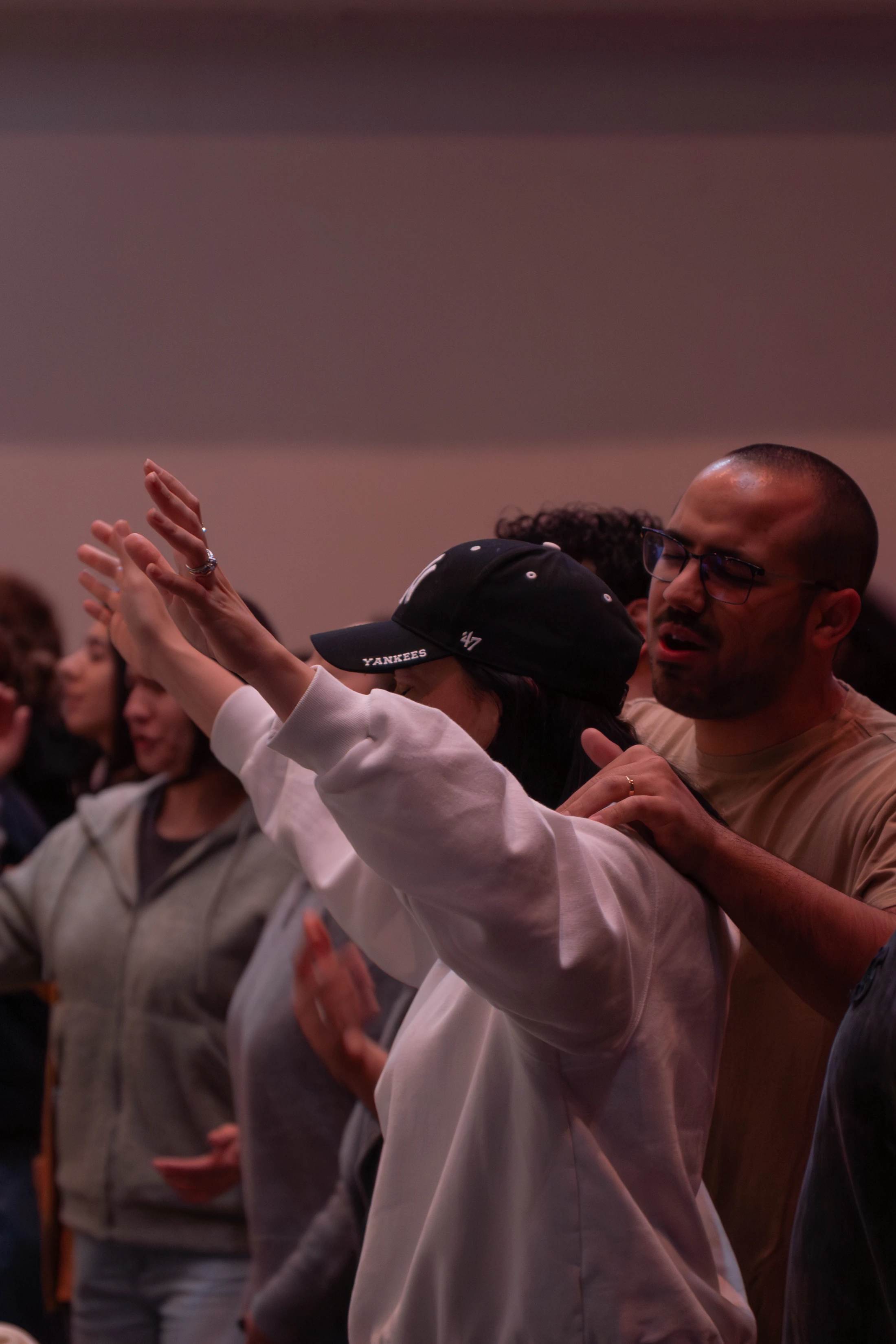 A woman in a baseball cap and a man beside her worship with eyes closed in a group. She has her hands raised, and he has a hand on her shoulder.