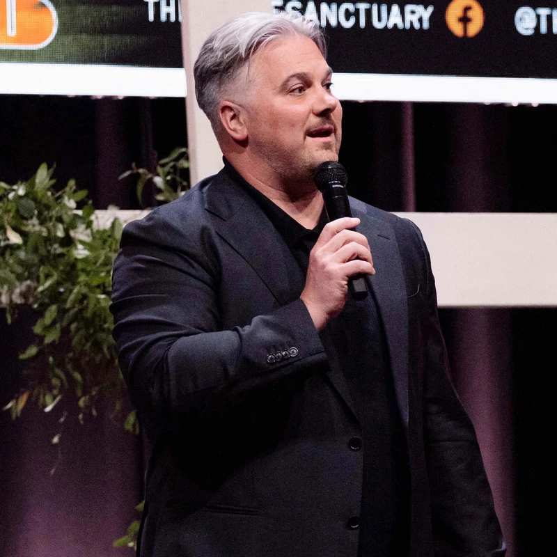 A man with grey hair wearing a black suit jacket holds a microphone and speaks on a stage. Behind him is a screen that says 'SANCTUARY'.