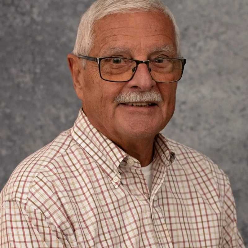 A headshot of an older man with white hair, a mustache, and glasses. He is wearing a plaid button-down shirt and smiling in front of a gray backdrop.