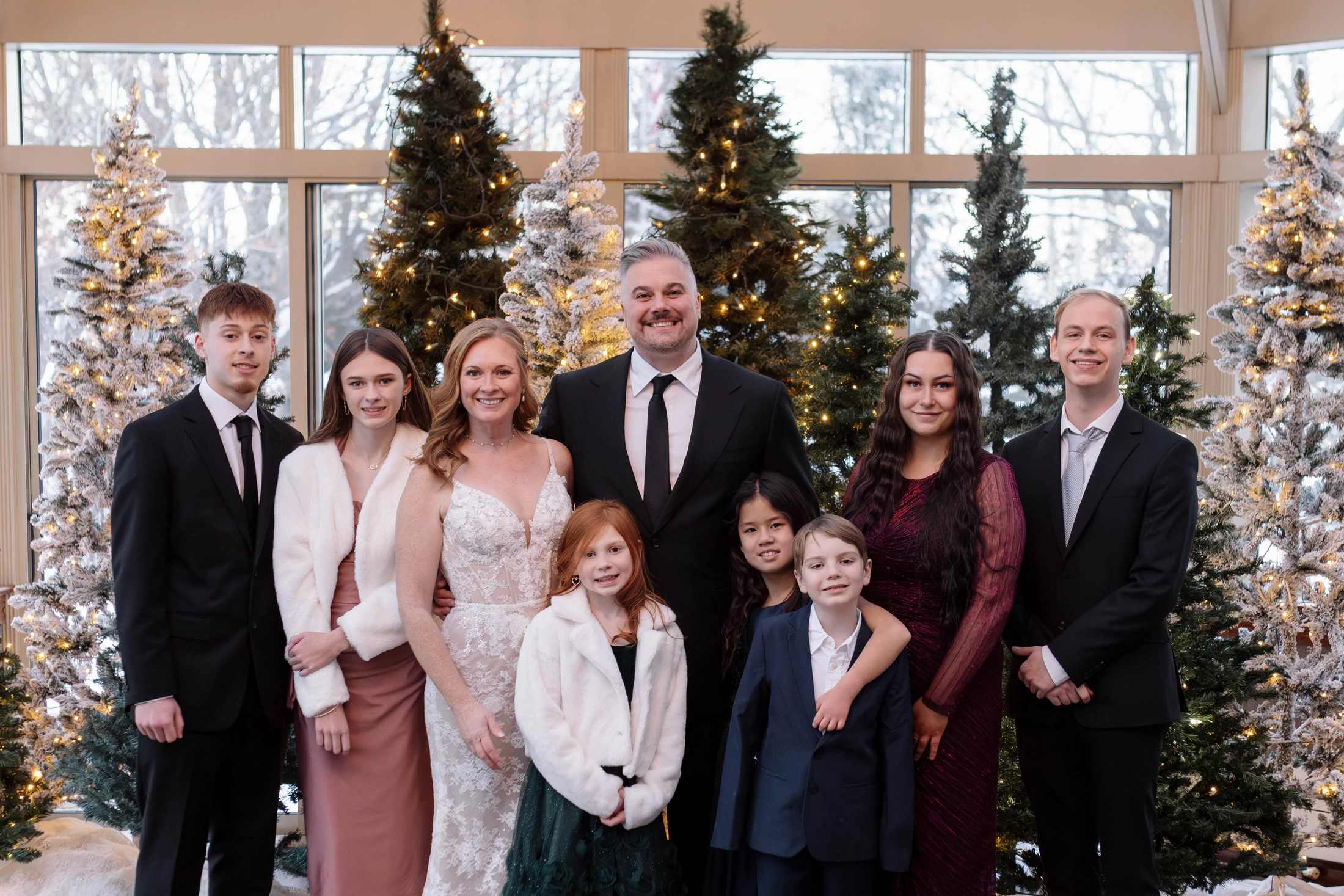 A newlywed couple smiles with their seven children for a formal portrait in front of several decorated Christmas trees at their winter wedding.