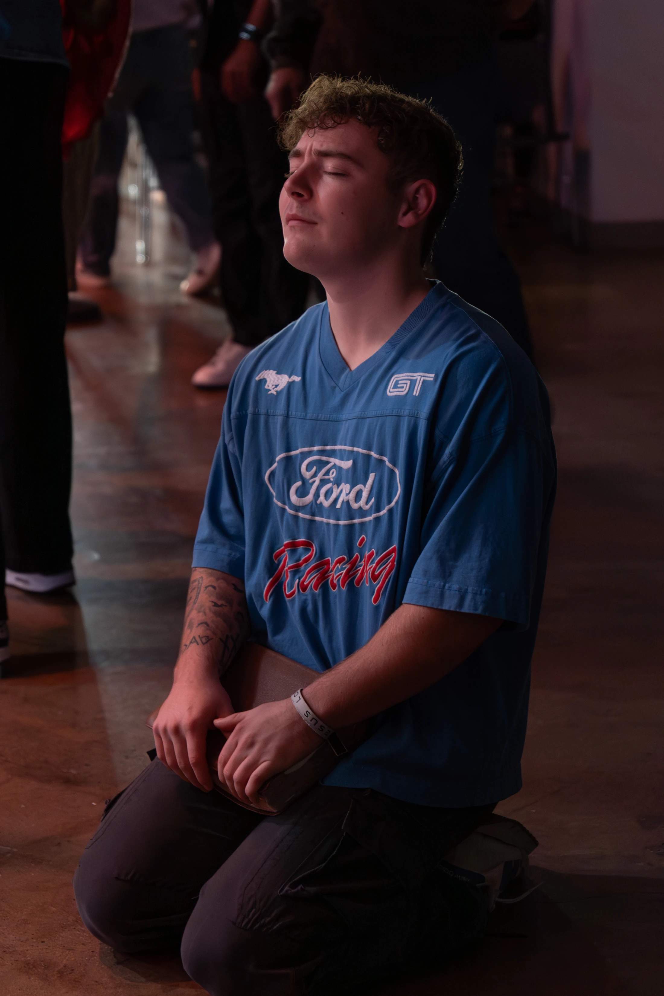 A young man with curly brown hair kneels on a wooden floor, his eyes closed in prayer. He is wearing a blue Ford Racing t-shirt and holding a book.