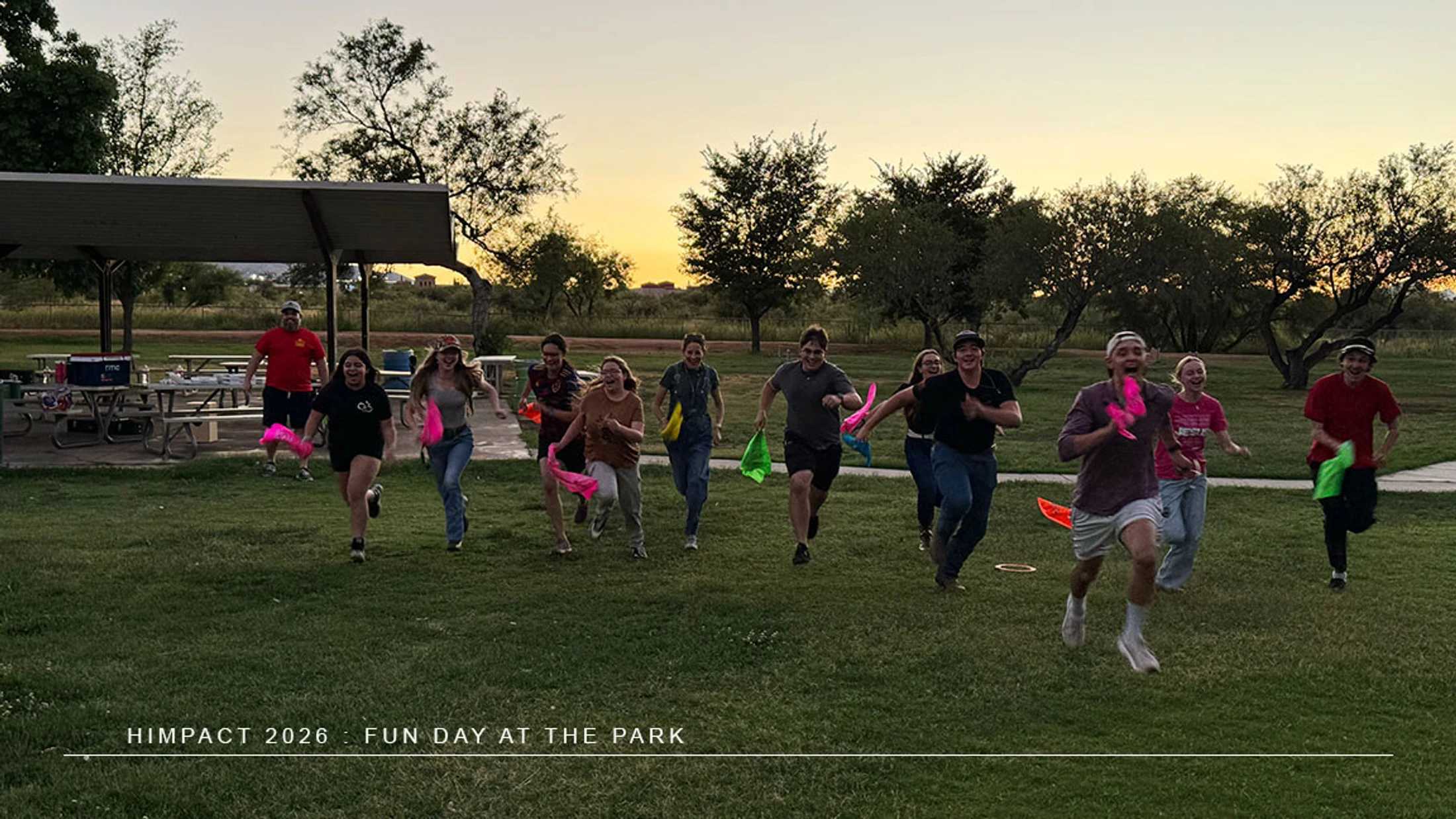A group of young people run joyfully across a grassy field at sunset, playing a game with colorful flags in a park with a picnic shelter in the background.