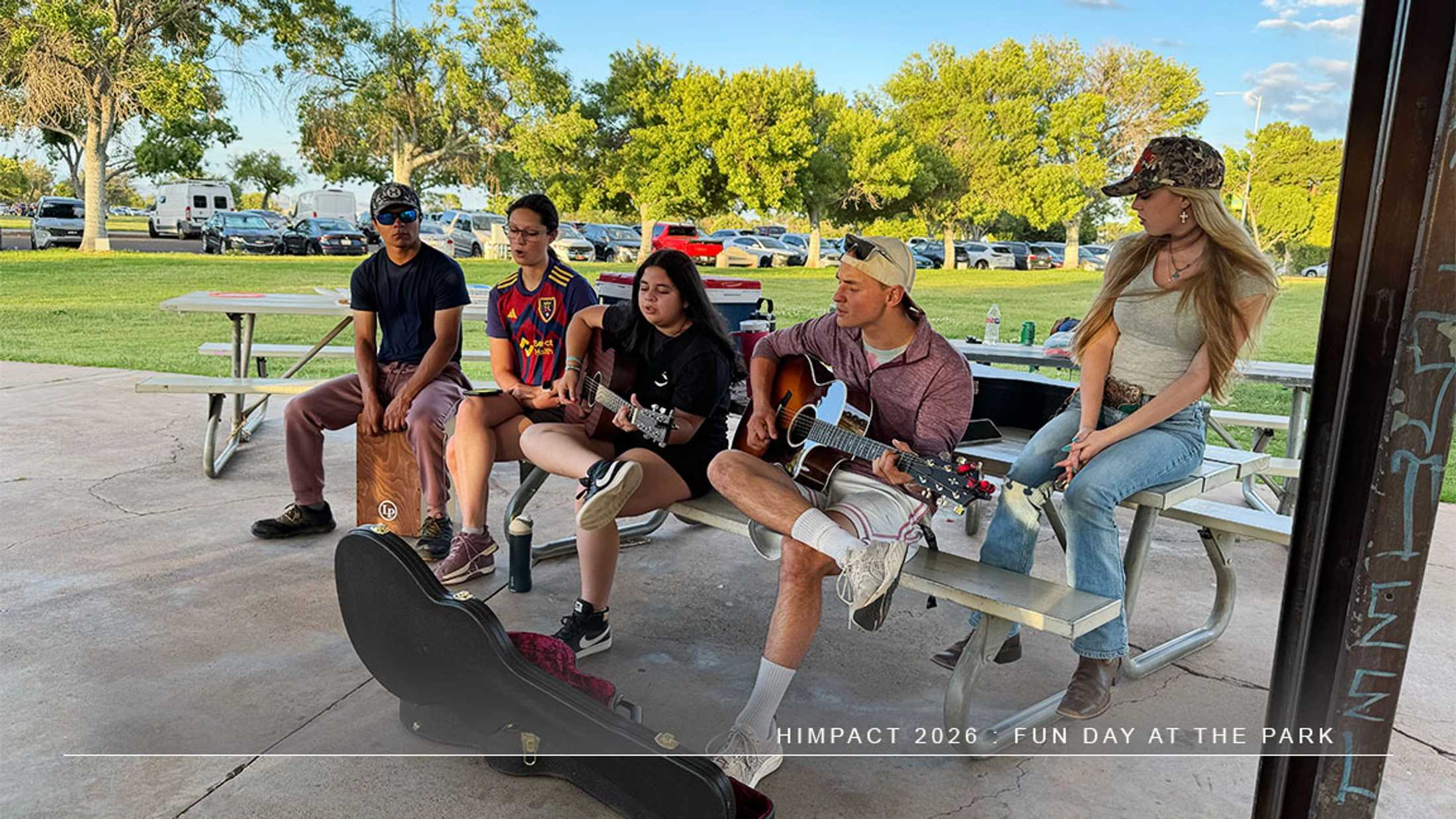 A group of five young adults sit at a picnic table in a park. Two are playing acoustic guitars and another plays a cajón drum.