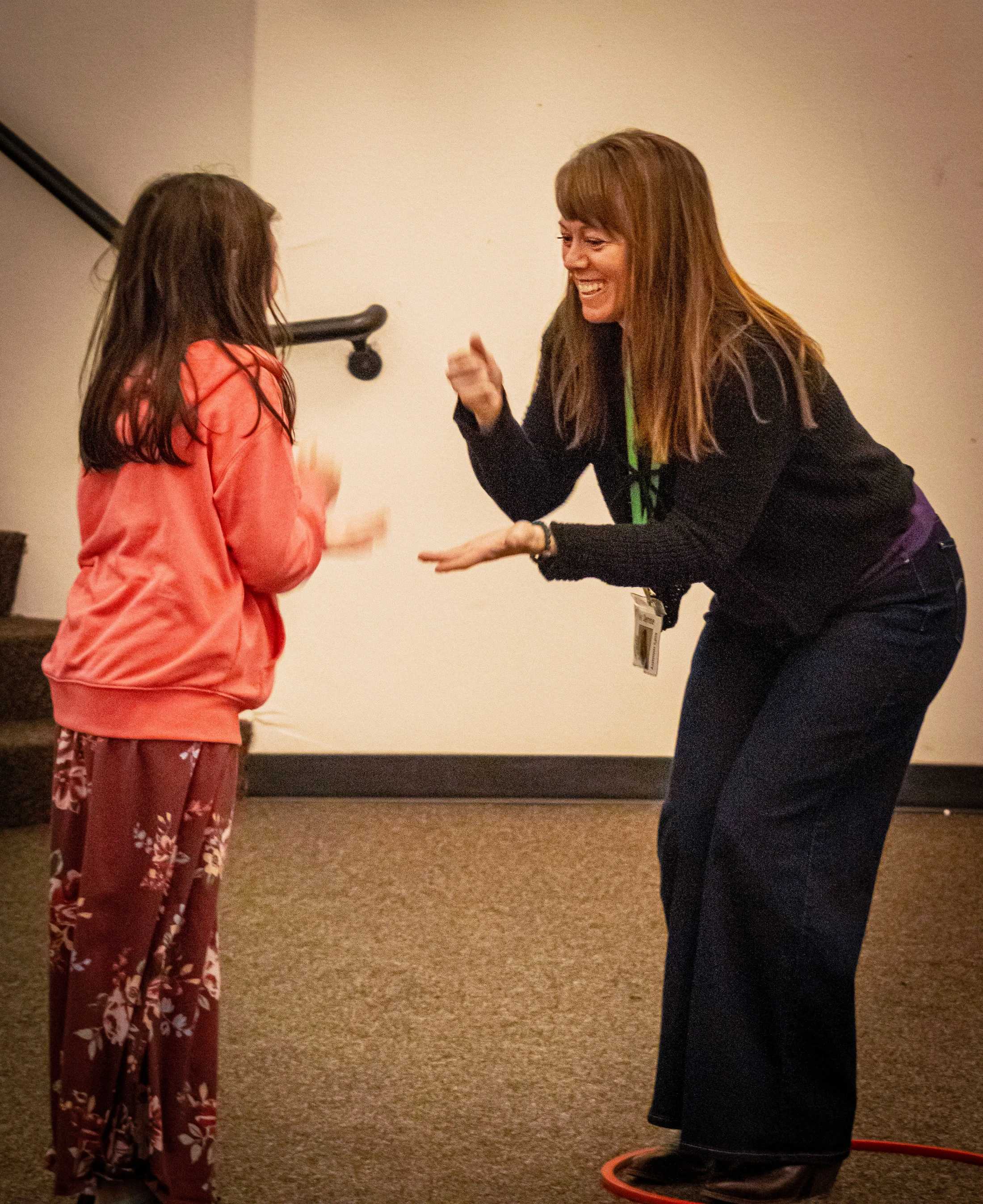 A smiling woman and a young girl with her back to the camera stand facing each other indoors, playing a hand-clapping game.