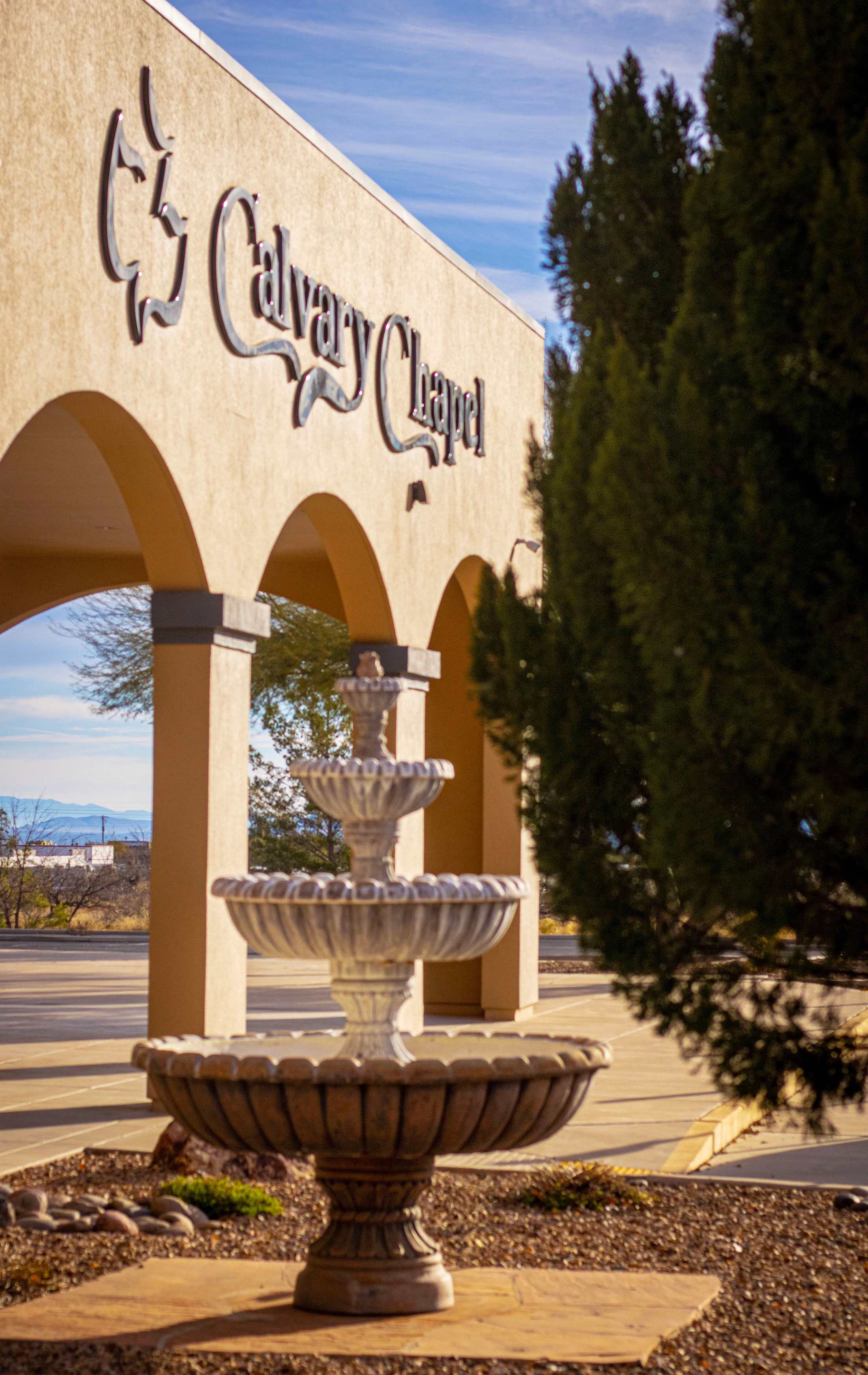 A three-tiered stone fountain sits in a rock garden in front of the arched entrance to Calvary Chapel under a blue sky.