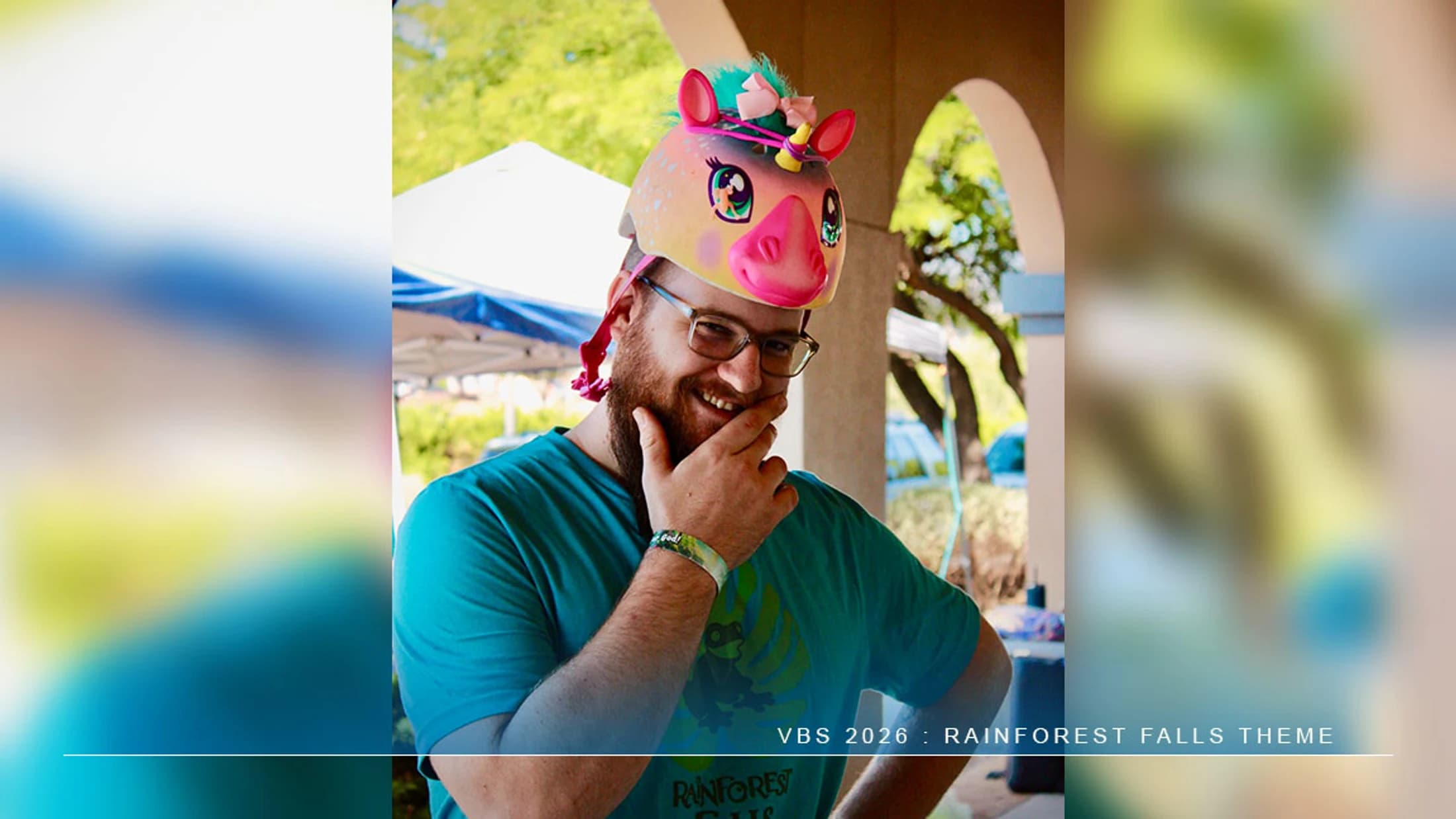 A smiling man with a beard and glasses wears a colorful, cartoon unicorn helmet and poses playfully with his hand on his chin.