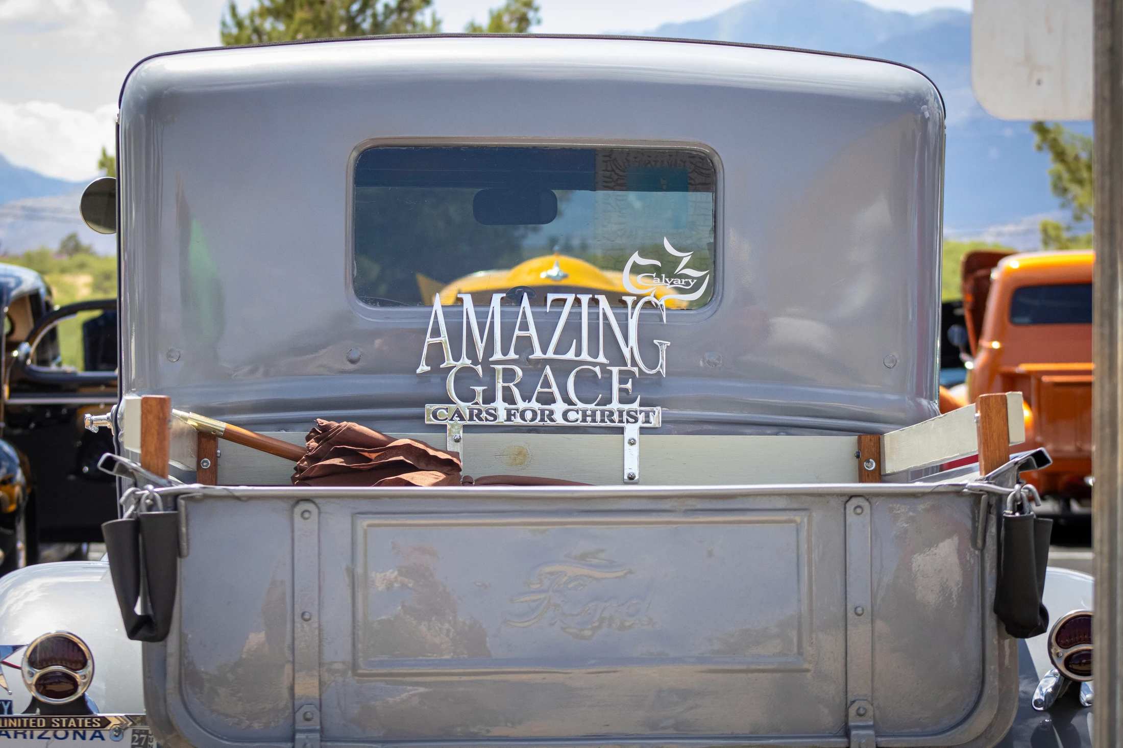 The back of a vintage gray Ford pickup truck with a chrome emblem that reads 'Amazing Grace, Cars for Christ'. A brown umbrella rests in the truck bed.