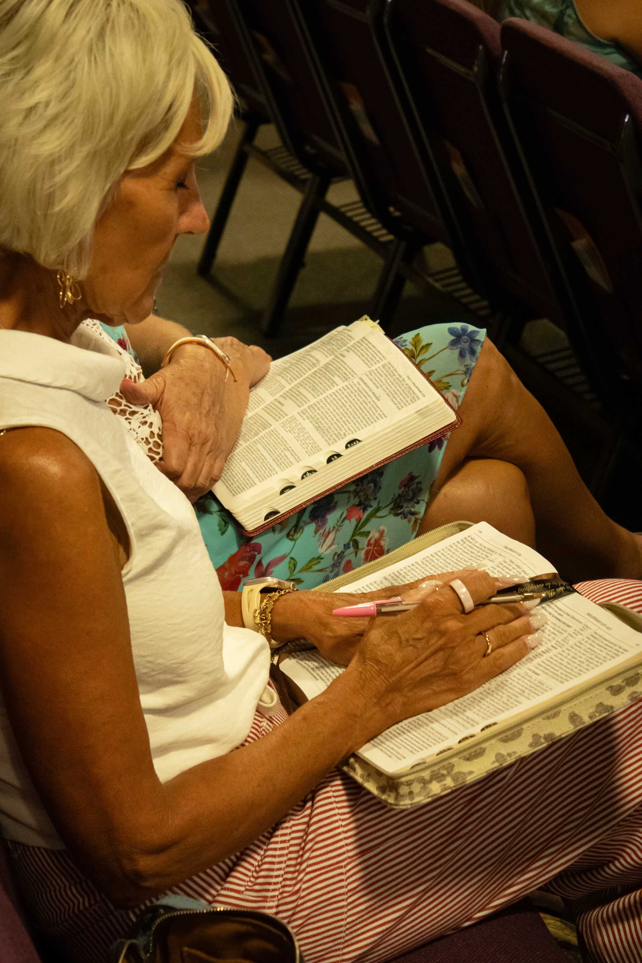 Two women sit side-by-side, each with an open Bible on their lap. The woman in the foreground has short blonde hair and holds a pen over her book.