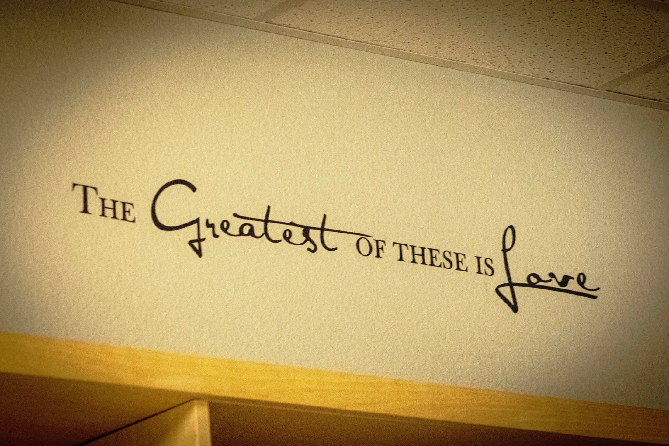 A wall decal in cursive and serif fonts reads, 'The Greatest of these is Love' on a warmly lit, beige textured wall above a wooden shelf.