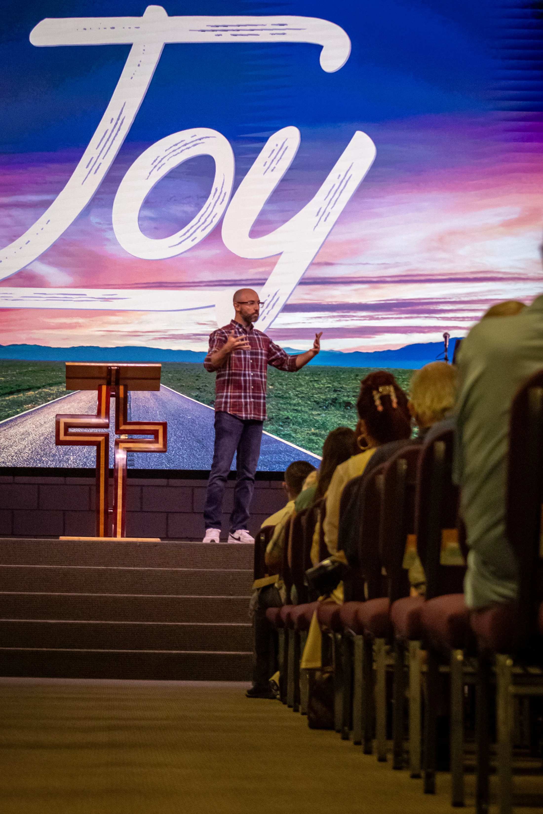 A man in a plaid shirt stands on a church stage, speaking to an audience. A screen behind him reads 'Joy' over a picture of a road and sunset.