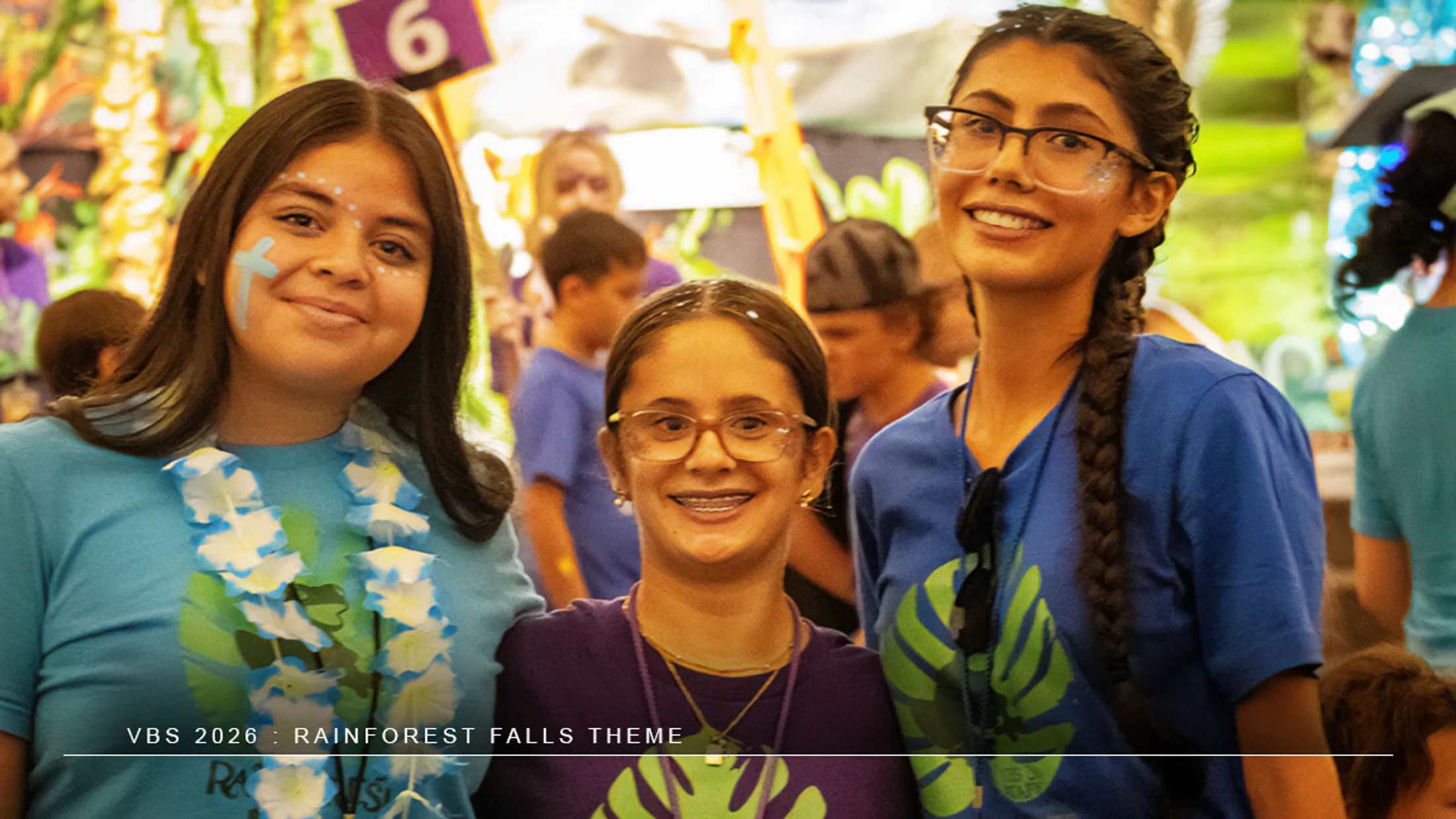Three young women smile for the camera at a VBS event with a rainforest theme. One wears face paint and a lei, while another has glitter on her face.