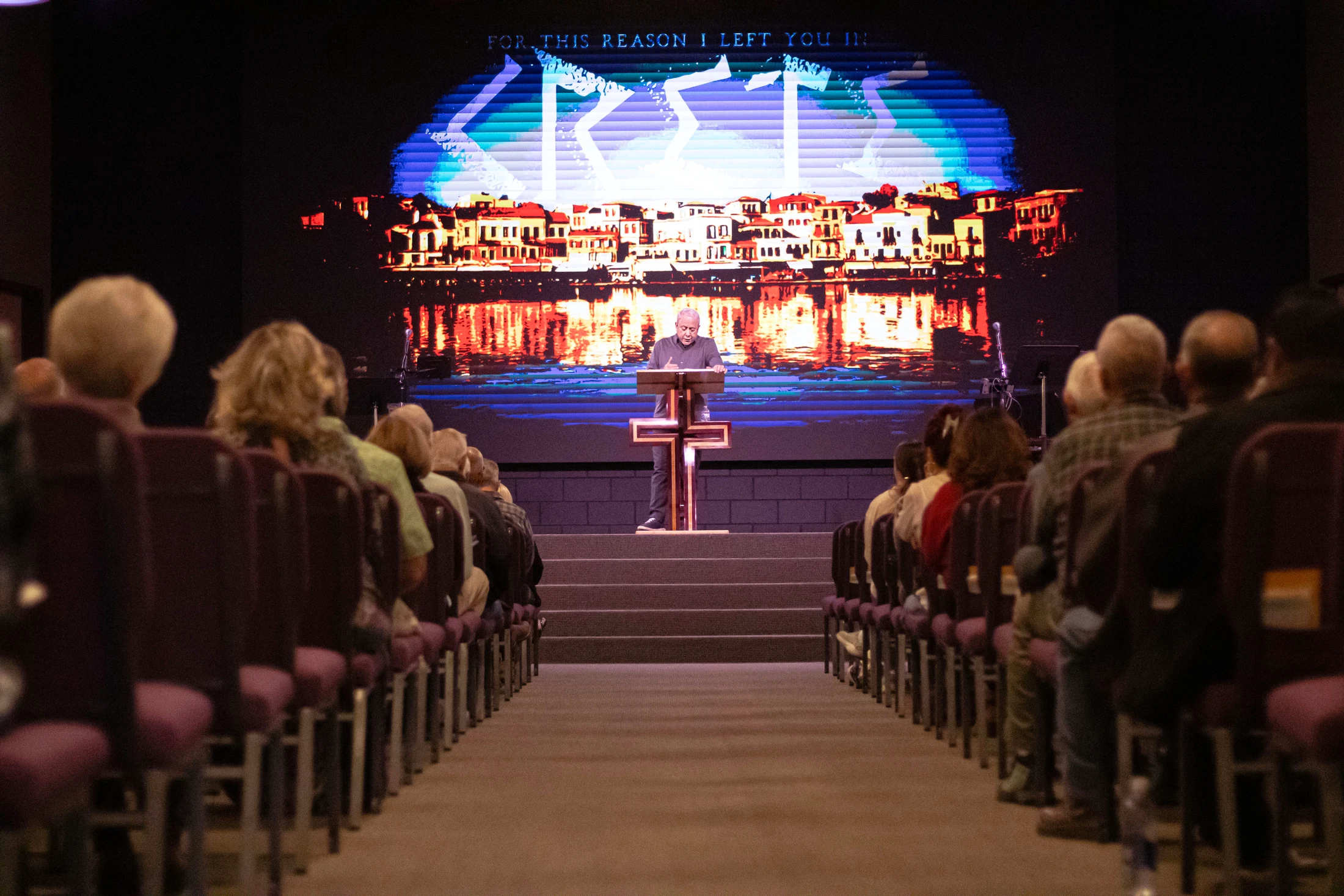 A view from the back of a church, looking down the center aisle at a pastor speaking from a podium on stage in front of a large screen.