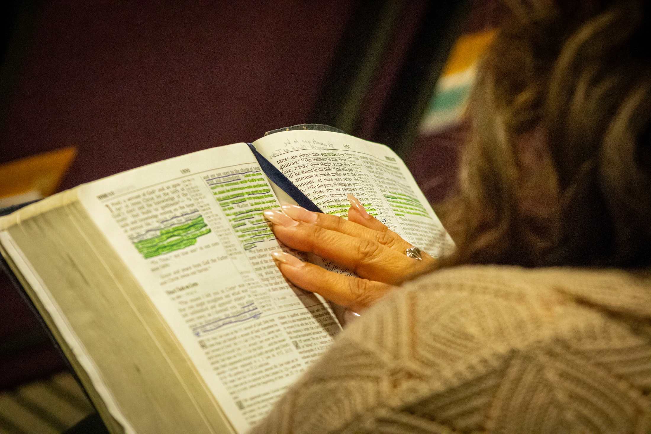 An over-the-shoulder view of a woman reading a Bible. Her hand, with manicured nails, rests on a page with highlighted and underlined passages.