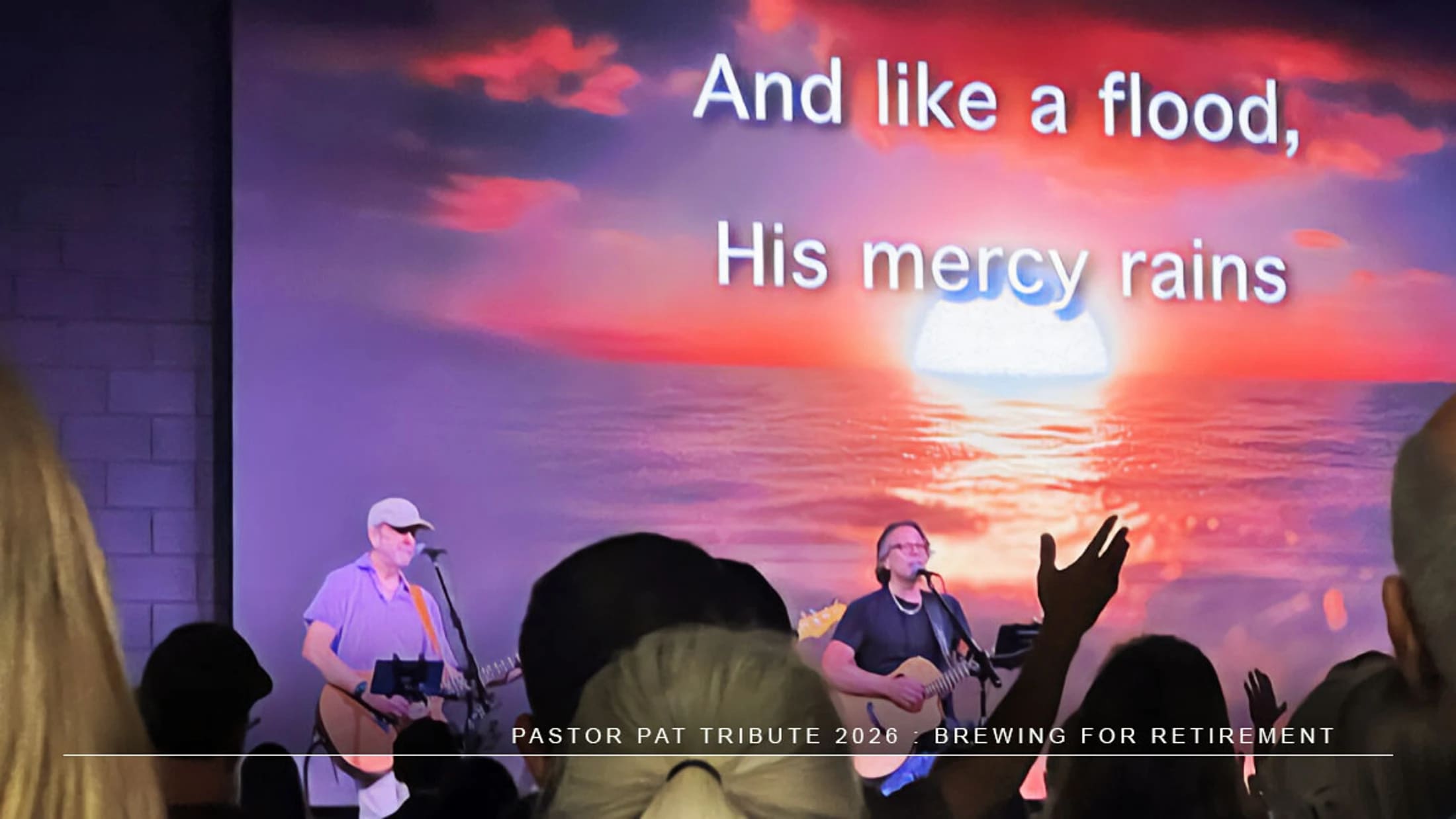 A band performs on stage for a pastor's tribute. A screen behind them shows a sunset over the ocean with the lyrics 'And like a flood, His mercy rains'.
