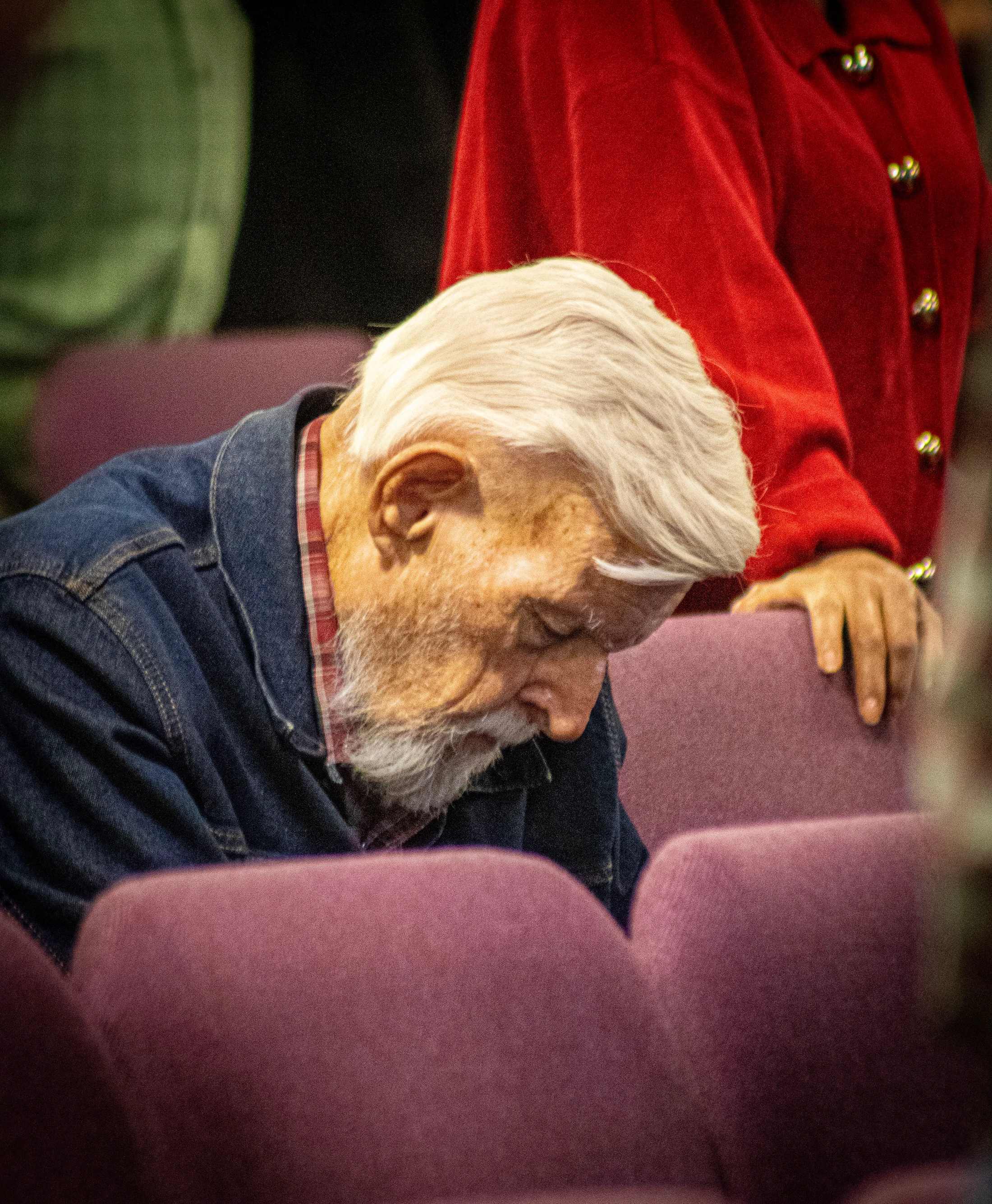 An elderly man with white hair and a beard, wearing a denim jacket, sits with his head bowed in a purple chair, seemingly in prayer or deep thought.