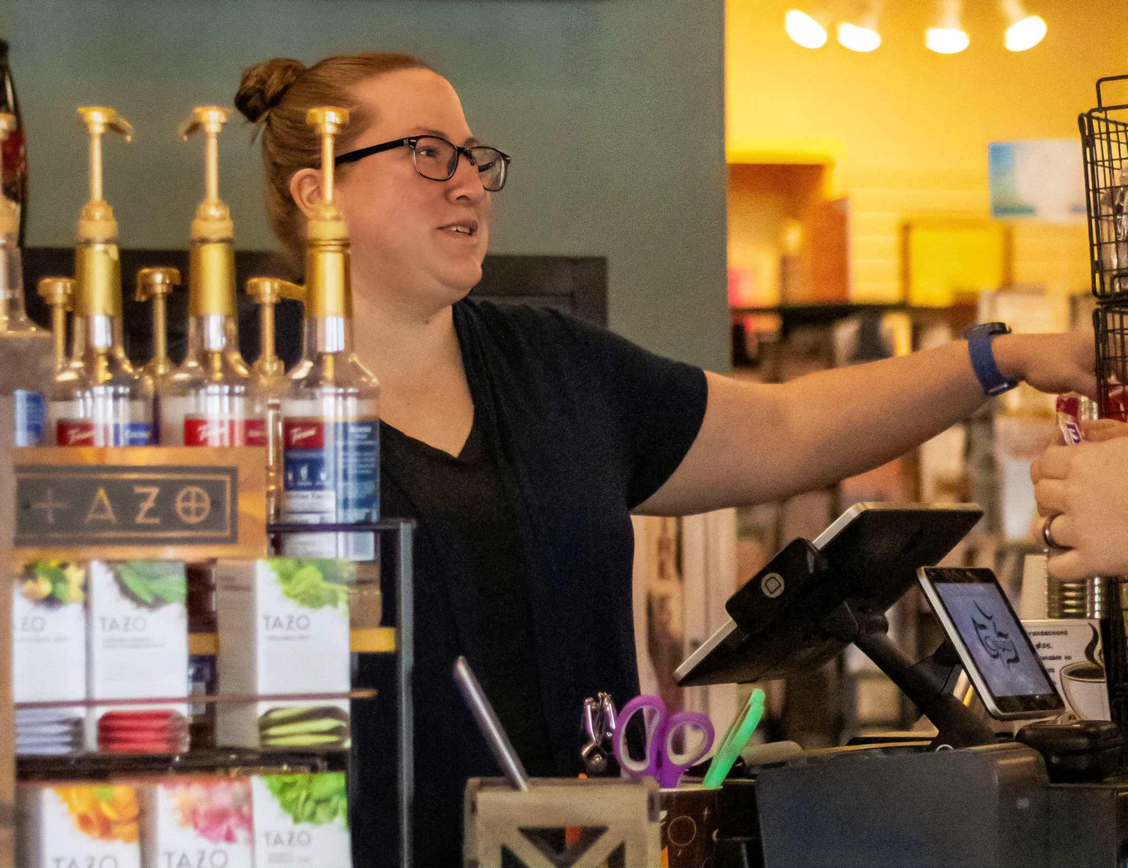 A female barista with glasses and her hair in a bun smiles while handing an item to a customer over a counter in a coffee shop.