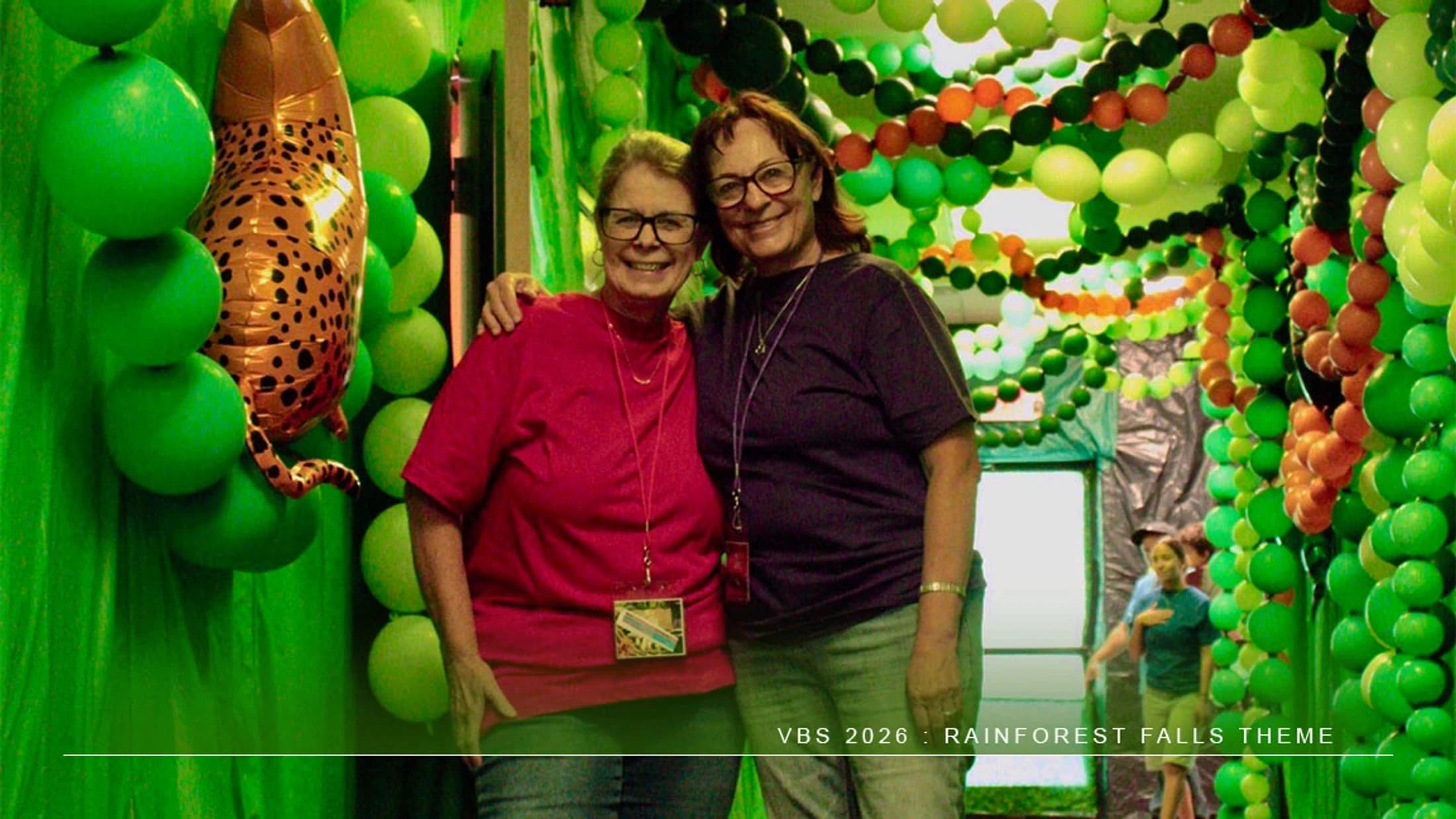 Two smiling women pose together in a hallway decorated with hundreds of green balloons and a leopard balloon for a 'Rainforest Falls' themed VBS.
