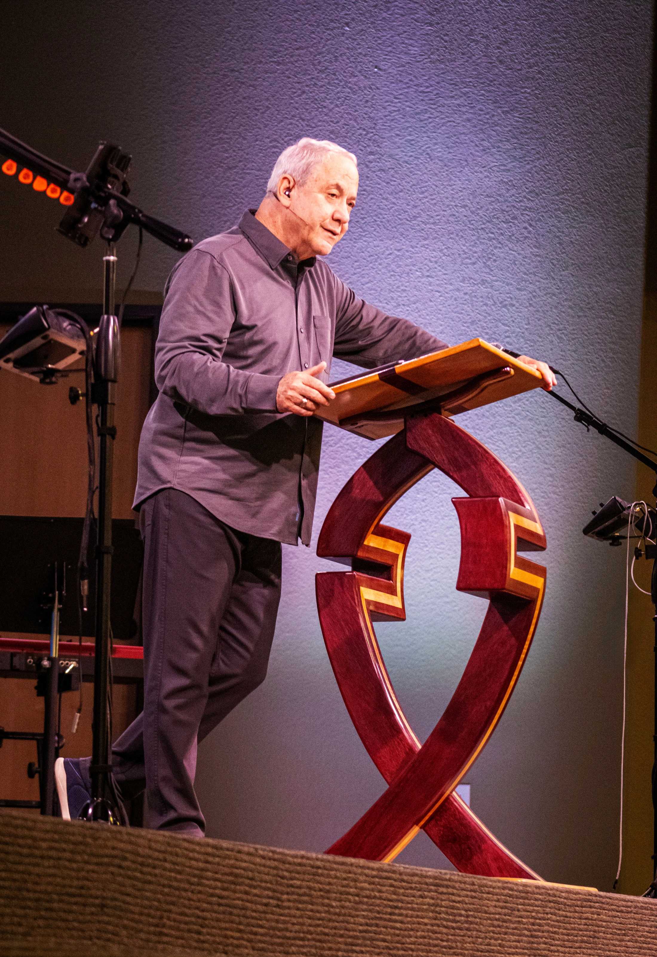 An older man with grey hair in a grey button-down shirt stands on a stage behind an ornate, modern wooden lectern while giving a presentation.