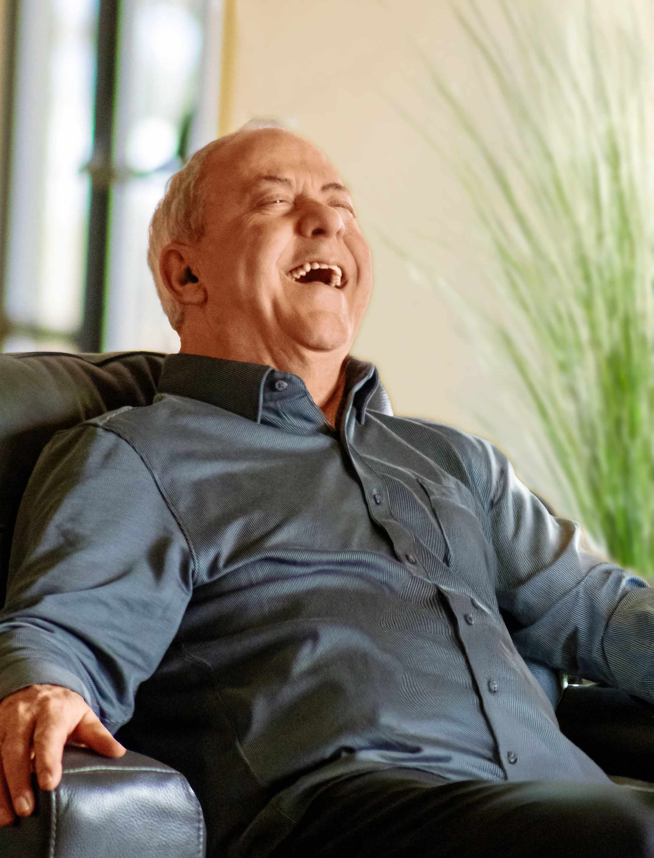 An older man with short gray hair sits back in a dark armchair, laughing heartily with his head tilted back and mouth open in a brightly lit room.