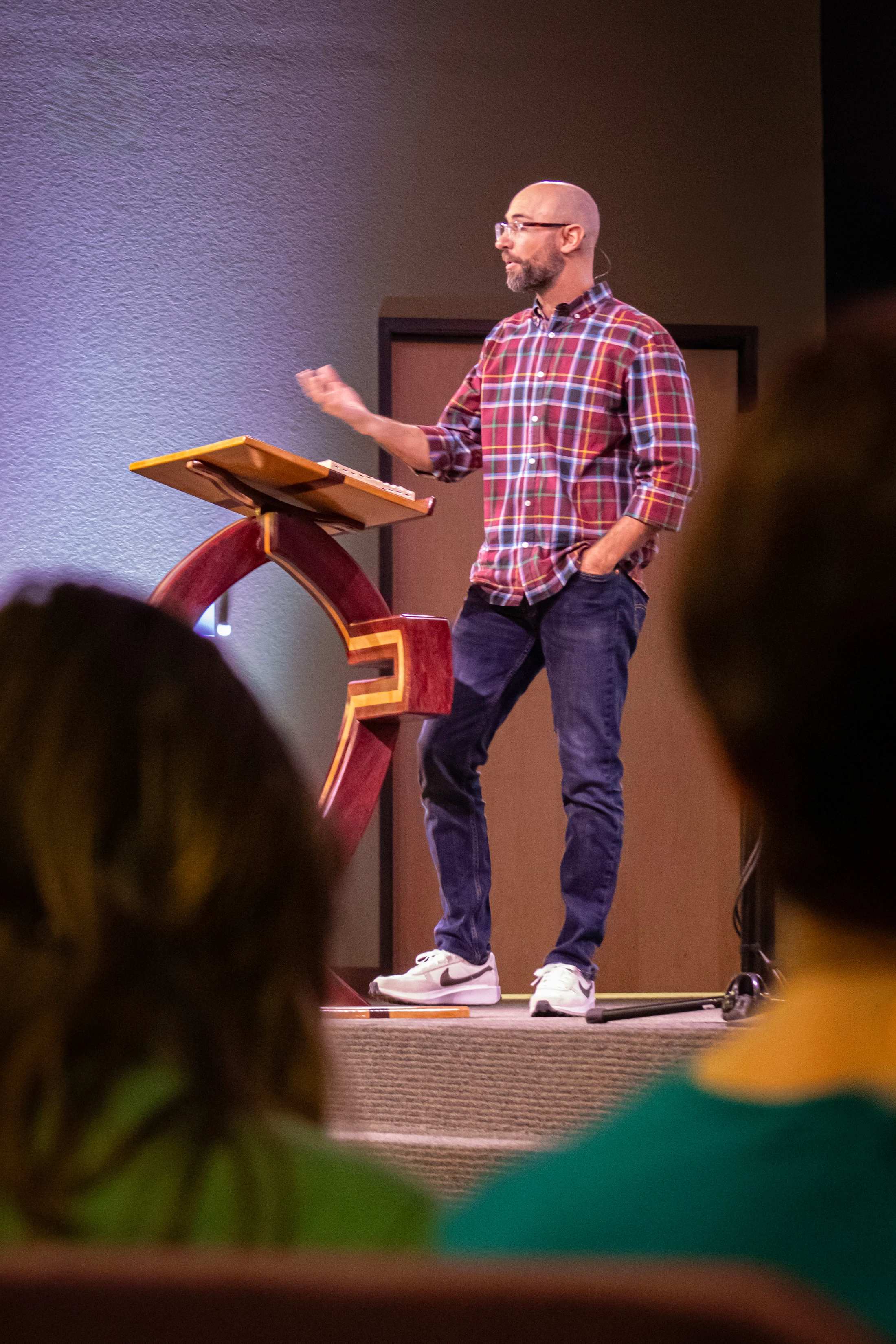 A bald man with glasses and a beard, wearing a plaid shirt and jeans, stands on a stage behind a wooden lectern, gesturing as he speaks to an audience.