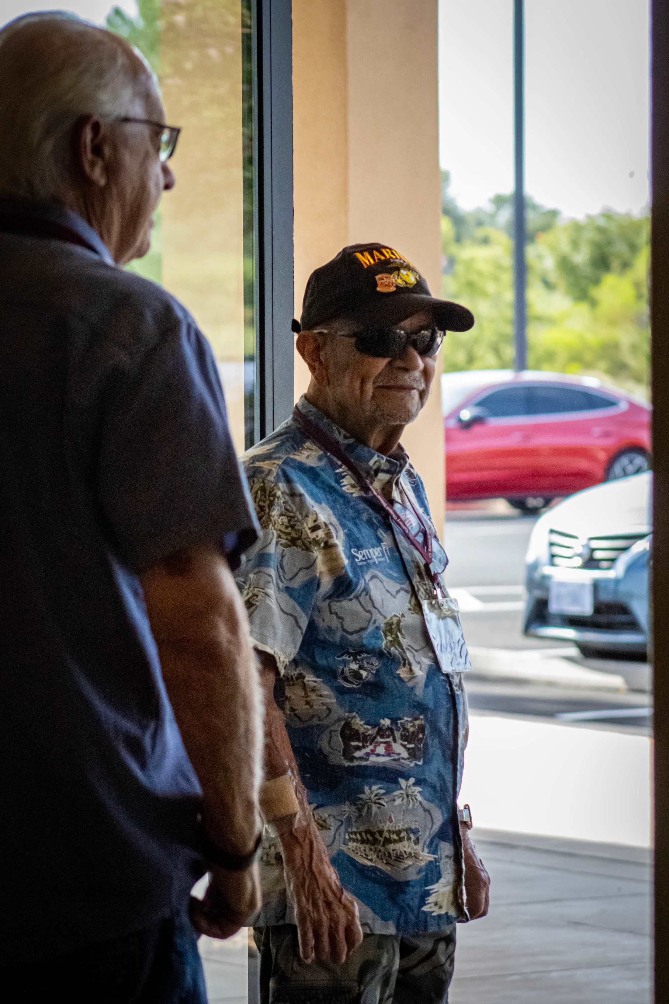 An elderly man in a Marines cap, sunglasses, and a military-themed Hawaiian shirt smiles while standing in a doorway, talking to another man.