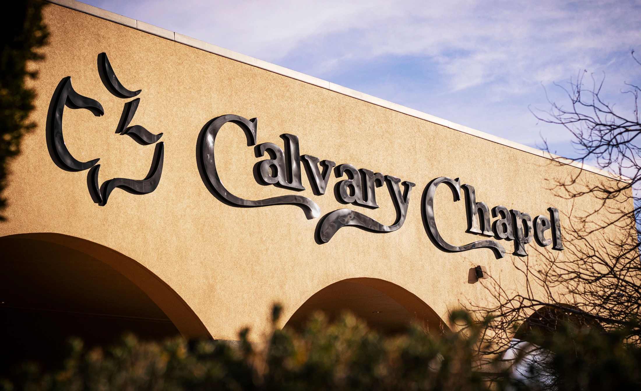 The black sign for Calvary Chapel, with its dove logo, is mounted on the tan stucco wall of the church building under a partly cloudy blue sky.